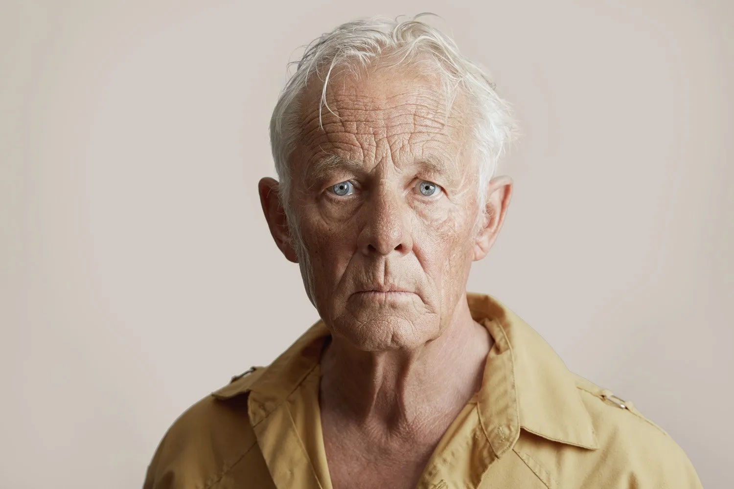 Retrato de un hombre mayor con cabello canoso y ojos azules, viste una camisa de color beige, fondo neutro.