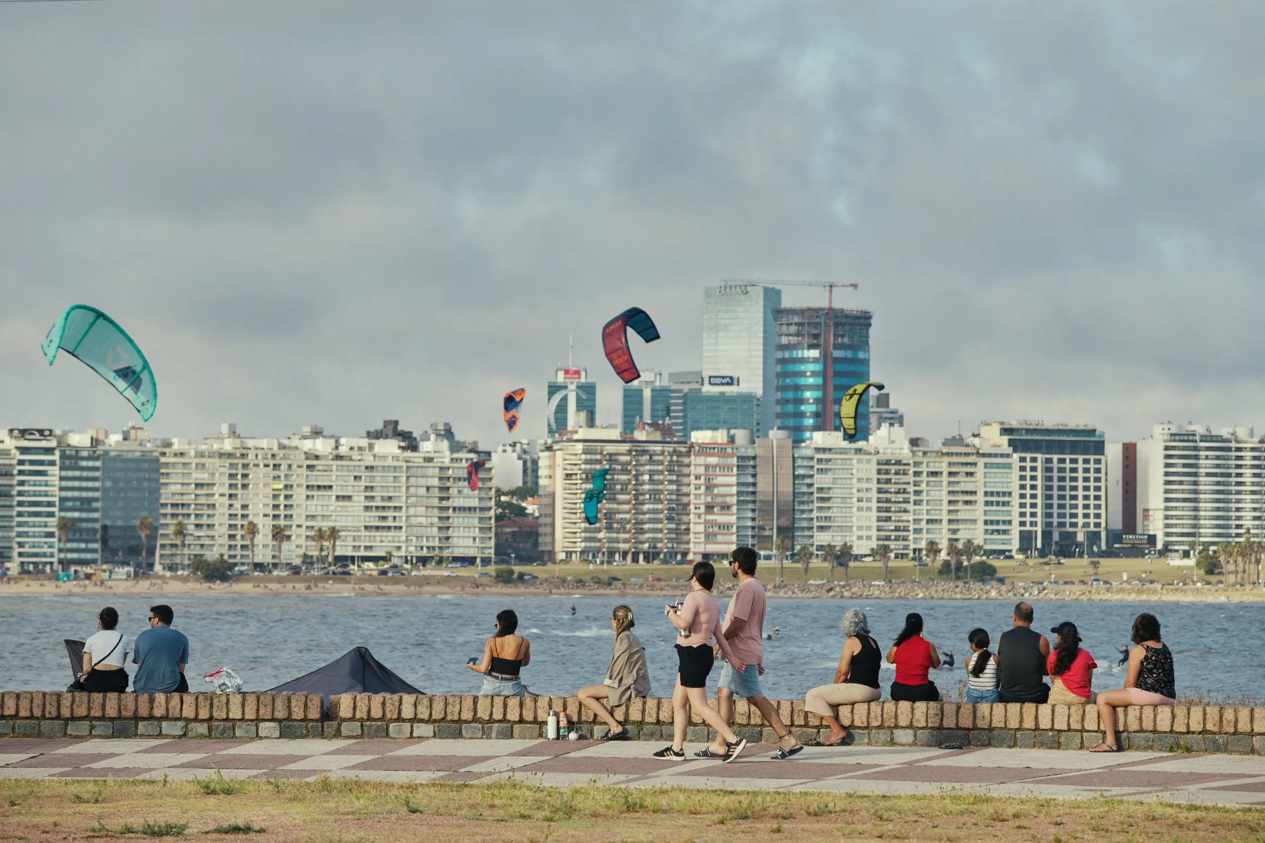 Gente sentada en el muro de la Rambla de Pocitos, con los kites sobre el agua y los edificios detrás
