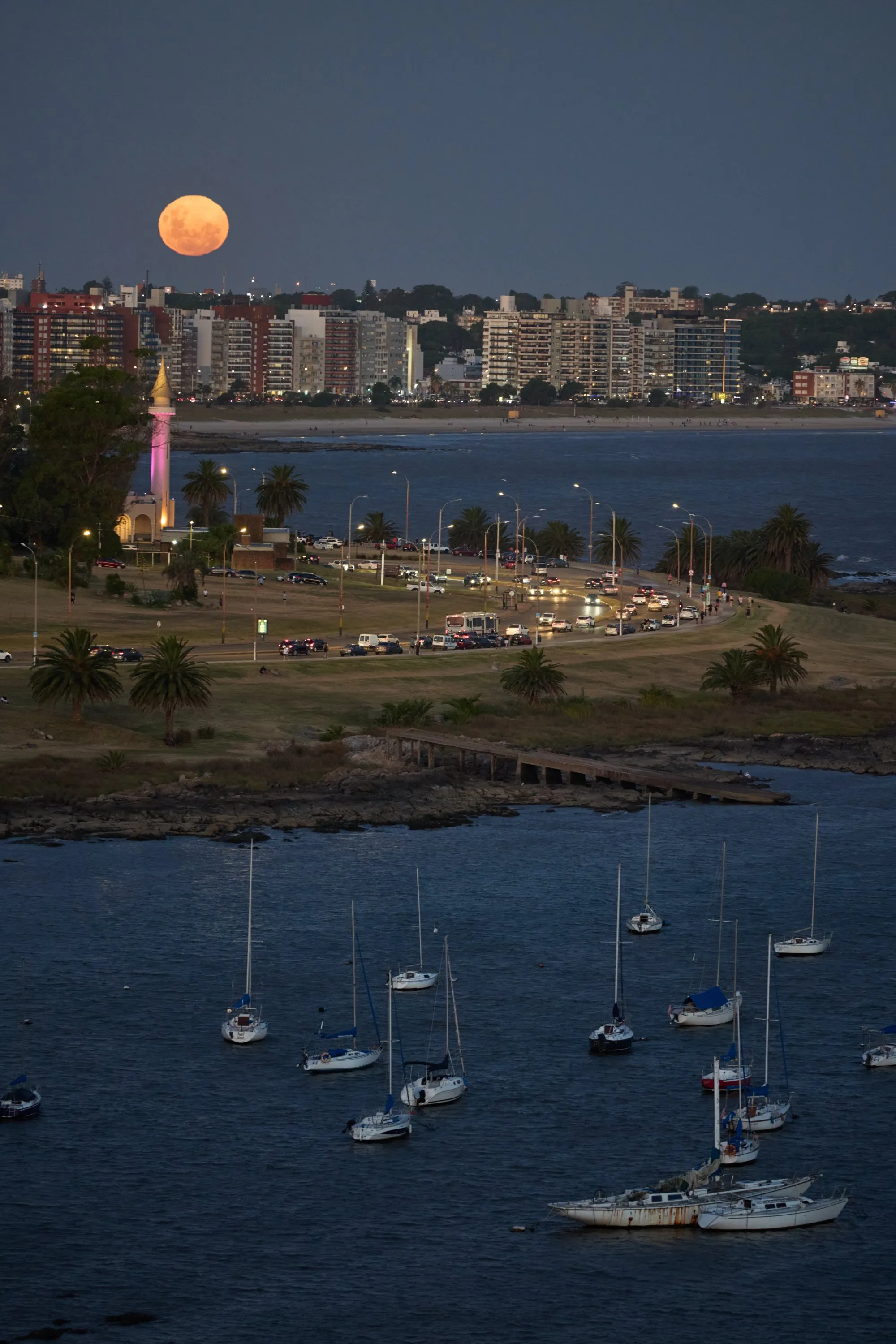 La Rambla de Montevideo a la altura del Buceo al anochecer, con el faro encendido y el tránsito sobre la costa