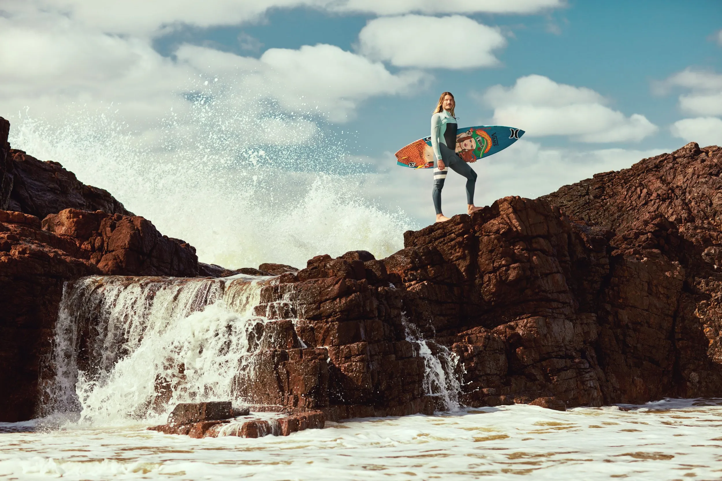 Persona con traje de neopreno en rocas junto al mar, sosteniendo una tabla de surf bajo el brazo, con olas rompiendo en el fondo y cielo parcialmente nublado.