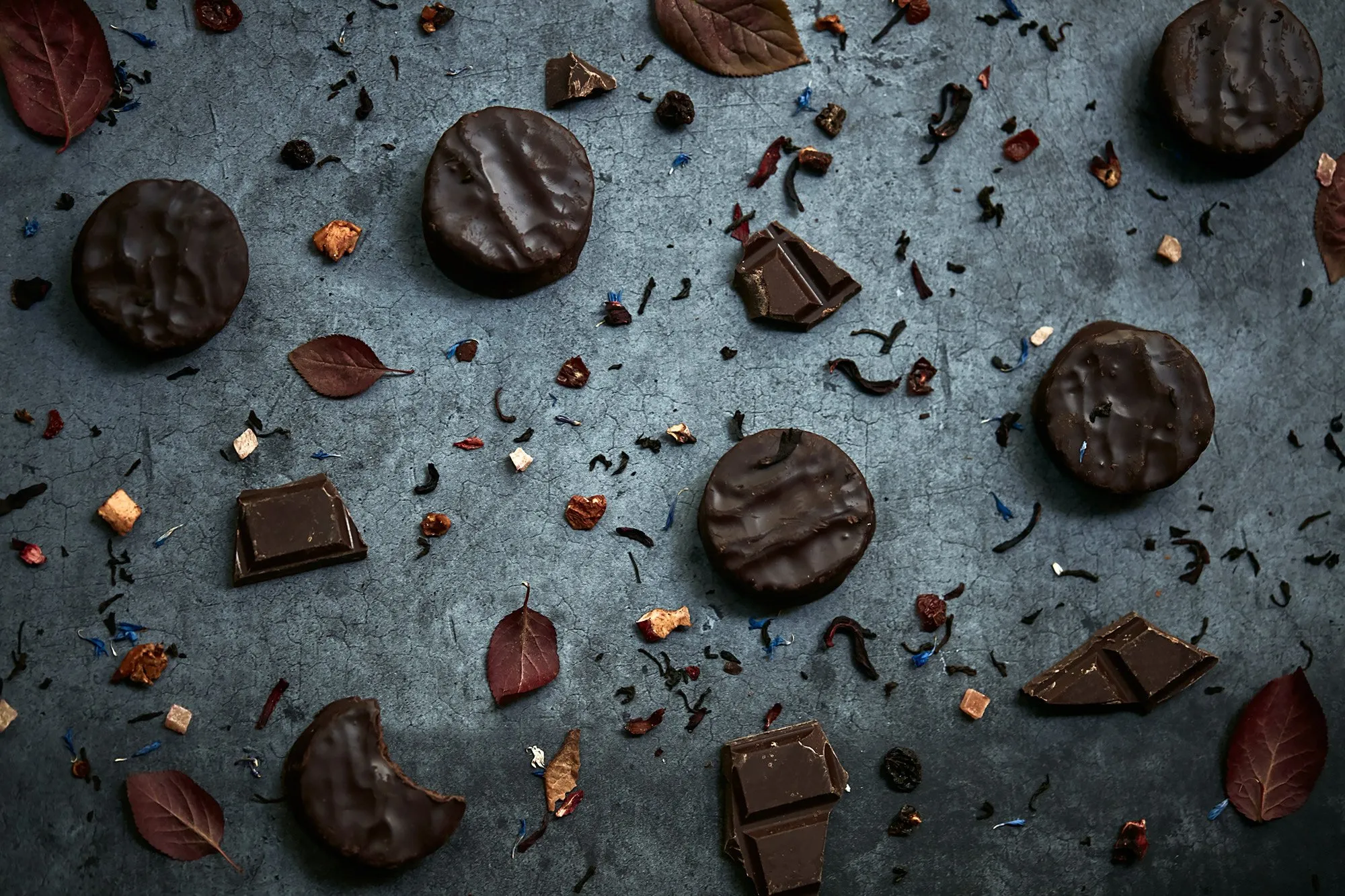 Varias piezas de chocolates envueltos, algunos rectangulares y otros redondos, con algunas hojas secas y pequeños trozos de ingredientes decorativos dispersos en un fondo oscuro.