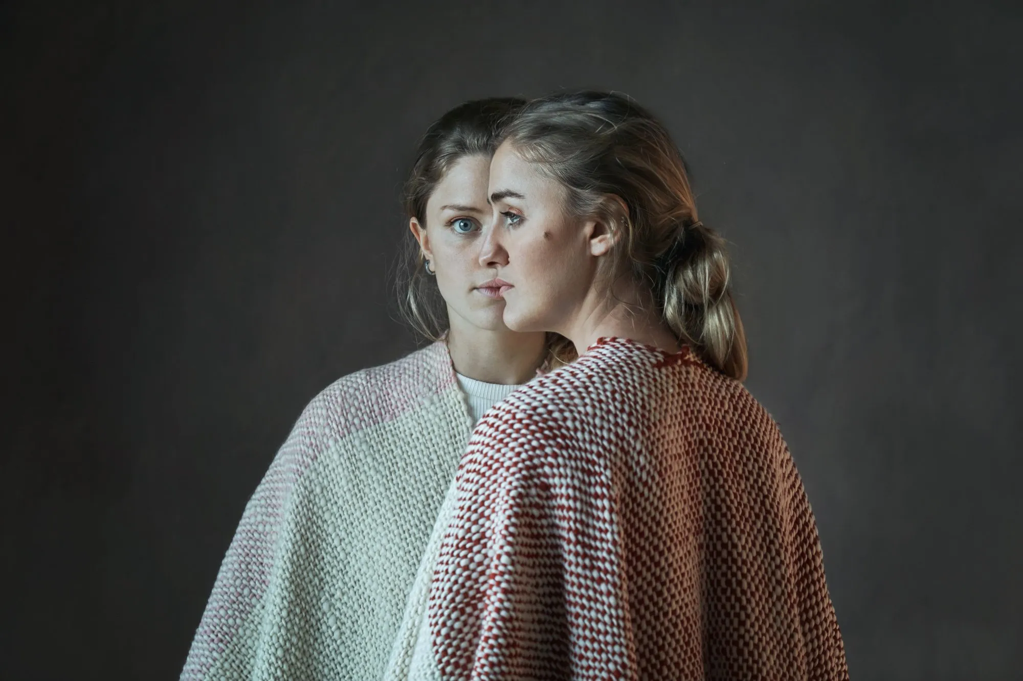 Dos mujeres con cabello rubio y liso, una frente a la otra, vestidas con suéteres de punto, en un fondo gris oscuro.