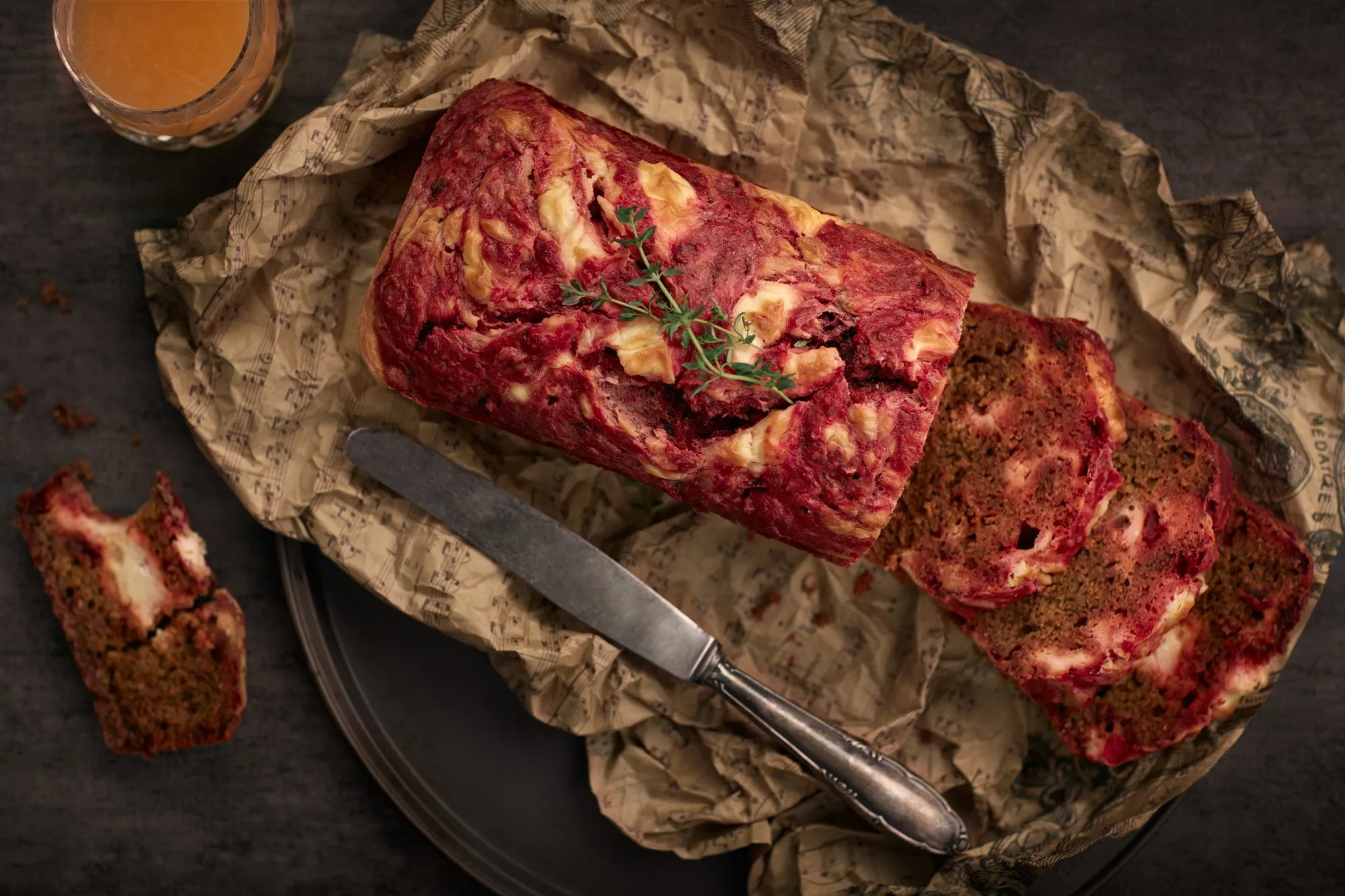 Pastel de carne sobre papel de periódico, acompañado de un cuchillo, con algunos rebanadas y una bebida en un vaso, en un fondo oscuro.