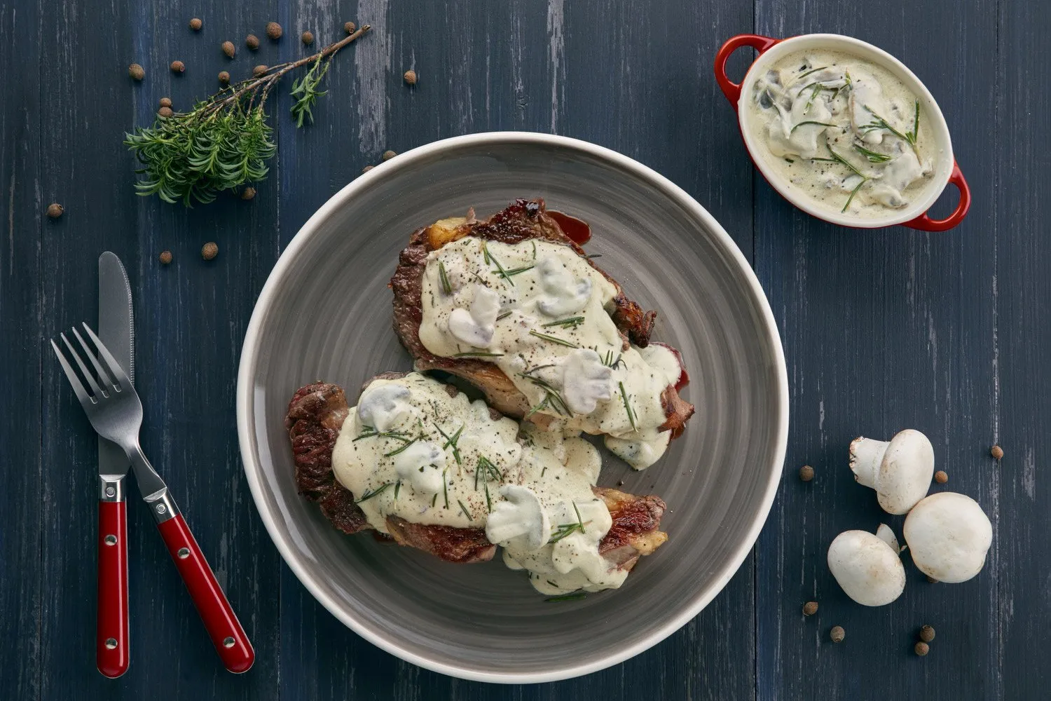 Carne con salsa bechamel y hierbas, acompañada de champiñones blancos y pimientas negras en una mesa de madera azul.