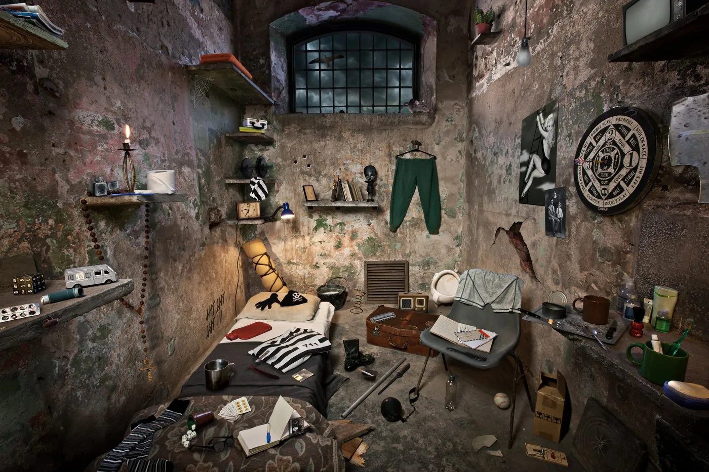 Una habitación desordenada con paredes de ladrillo expuesto y algunos objetos dispersos, incluyendo una cama, una silla, estanterías con libros y objetos, ropa colgada, y varias decoraciones en las paredes, en un ambiente con estilo descuidado y enve