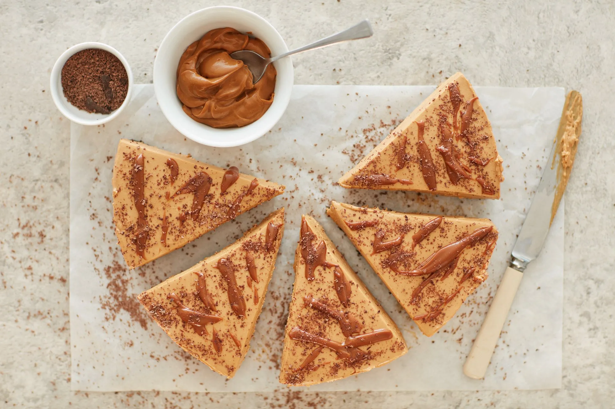 Seis rebanadas de pastel de caramelo con chocolate y chocolate en polvo en un papel encerado, con jarabe de chocolate en la parte superior, y dos recipientes con crema de manjar y cacao en polvo al lado.