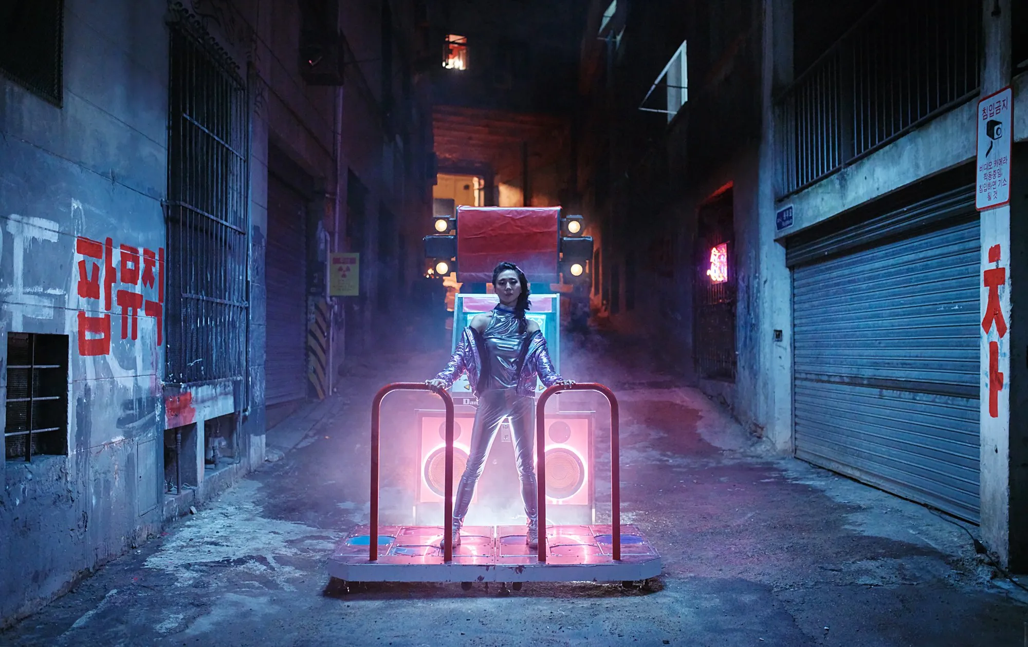 Mujer vestida con ropa futurista de pie sobre una plataforma en un callejón oscuro con neón y humo, con altavoces y una estructura de carga frente a ella.