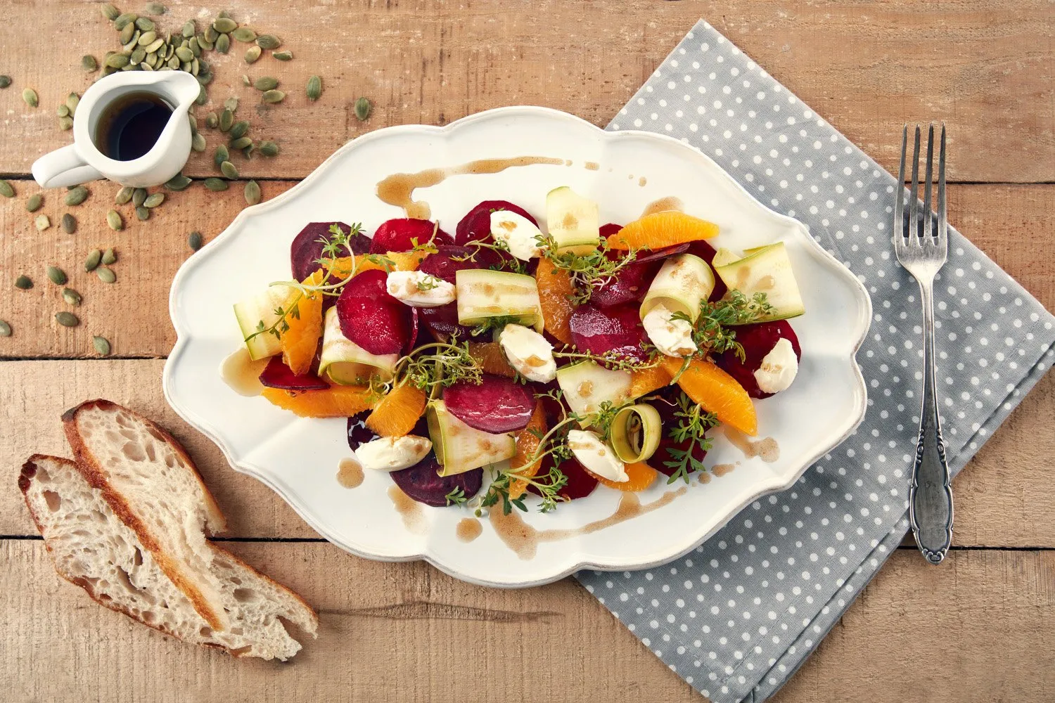 Ensalada de remolacha, naranja, pepino y queso en un plato ovalado blanco, con pan a un lado, sobre mesa de madera con servilleta de lino azul con puntos blancos y tenedor a la derecha.