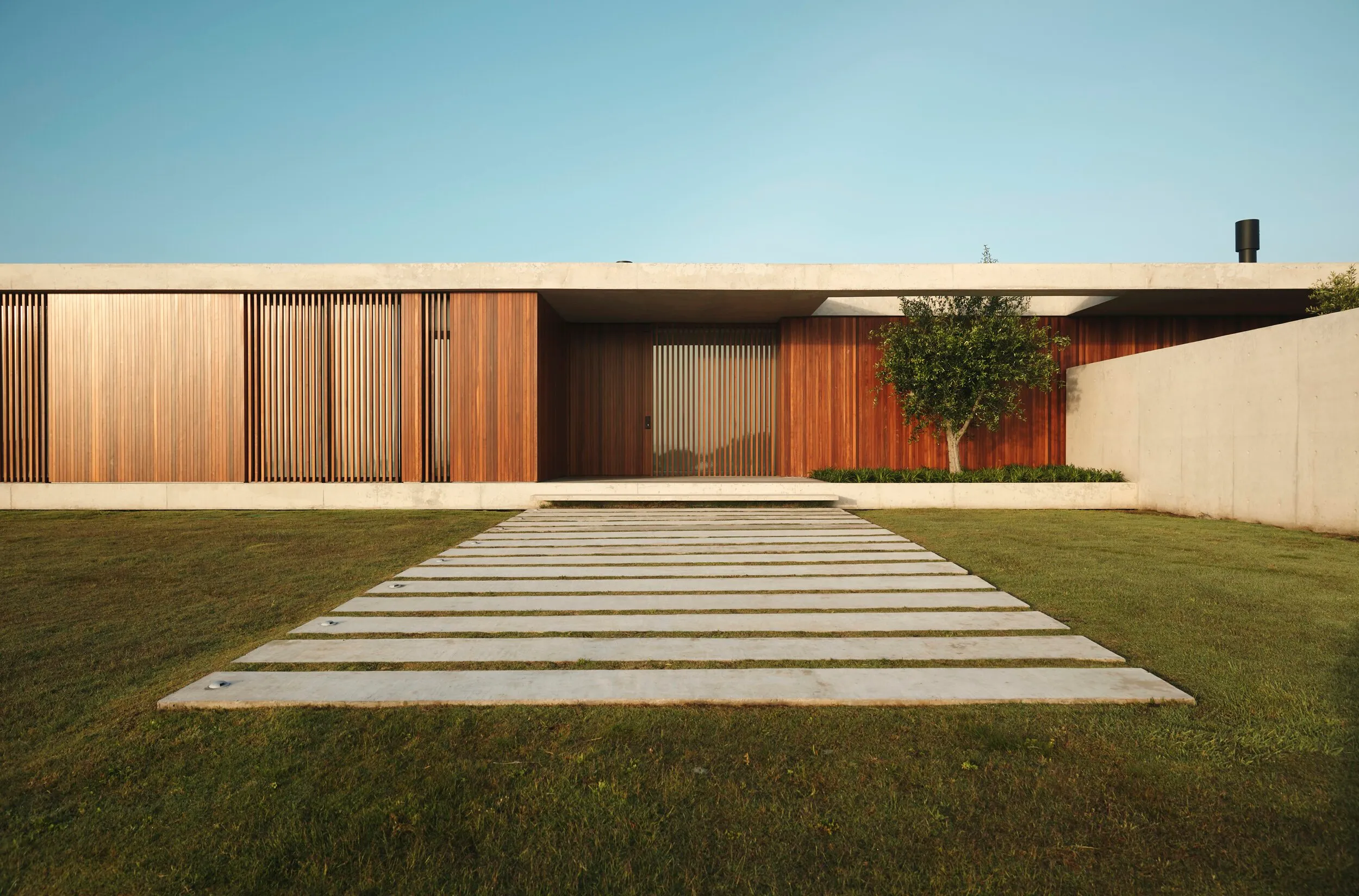 Casa moderna con fachada de madera y muro de cemento, camino de piedra en césped y árbol junto a la entrada.