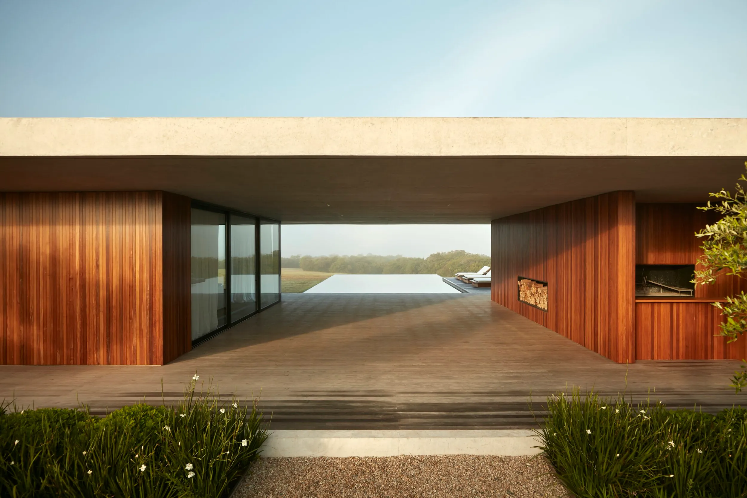 Vista frontal de una casa moderna con estructura de concreto y paredes de madera, vista de una piscina y sillas en el fondo, y áreas verdes en primer plano.