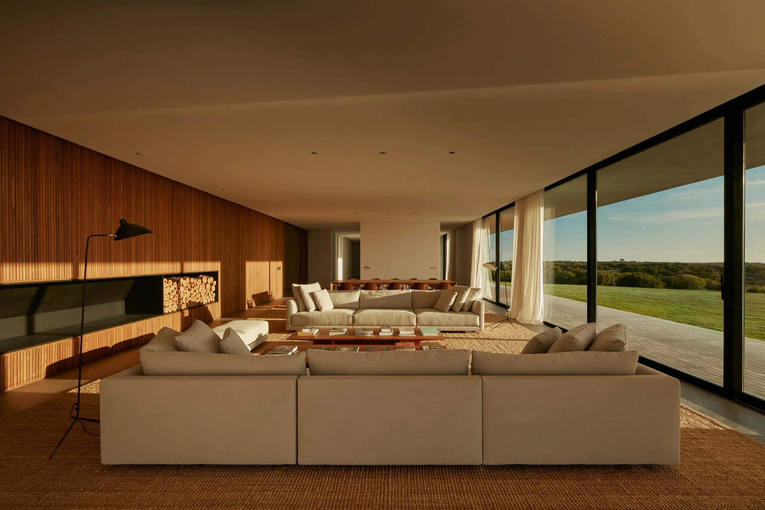Sala moderna con grandes ventanales, sillones beige, mesa de centro de madera, lámpara de piso negra, pared de madera y vista a un campo verde.