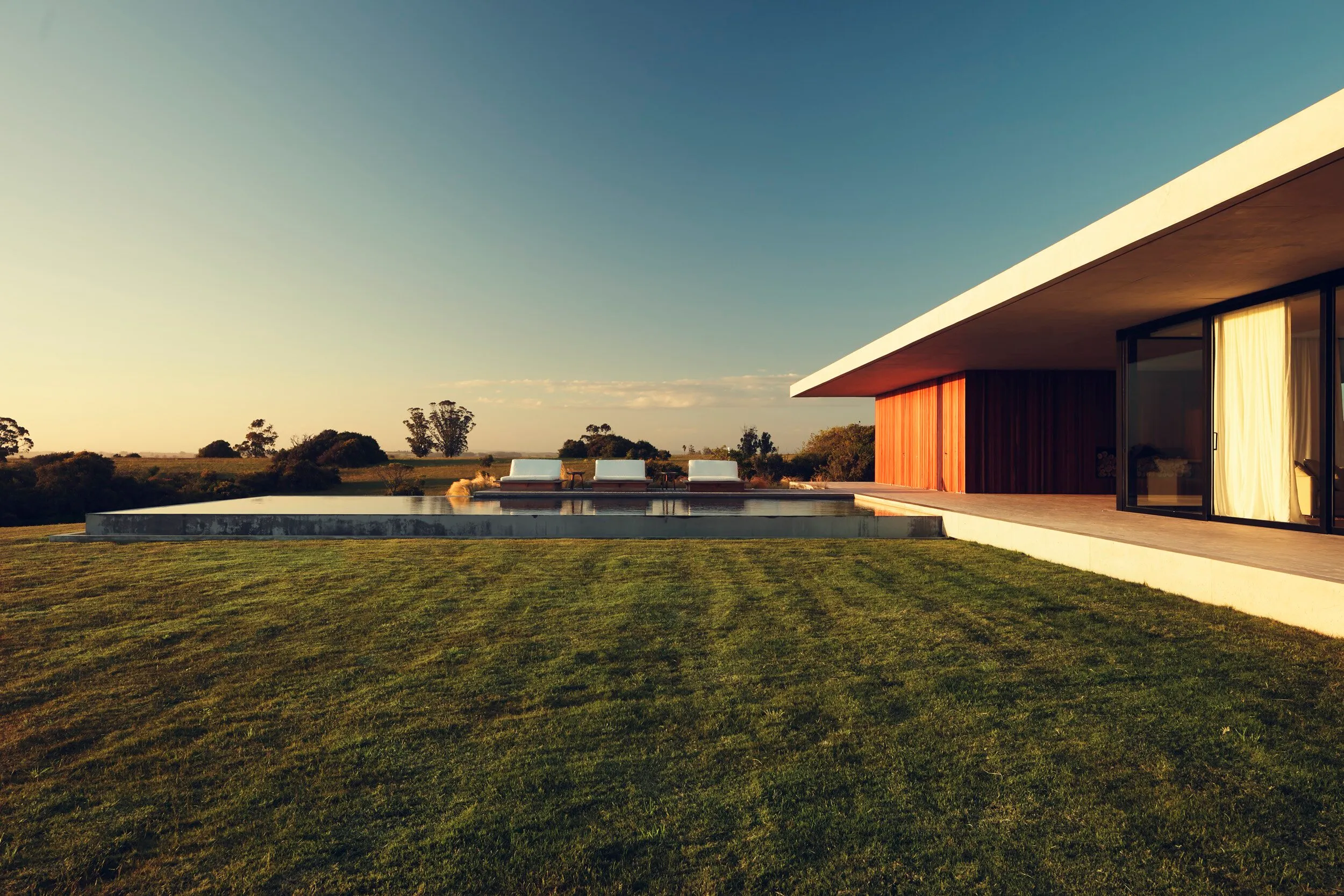 Casa moderna de un piso con fachada de madera y grandes ventanas, piscina en el jardín y sillas en el borde, en un entorno rural con césped y árboles
