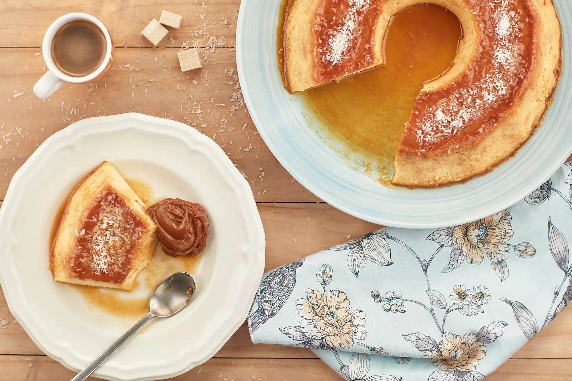 Un flan de caramelo en un plato, servido con una porción cortada y una porción de dulce de leche. También hay una taza de café y algunos cubos de azúcar en una mesa de madera decorada con un mantel con estampado de flores.
