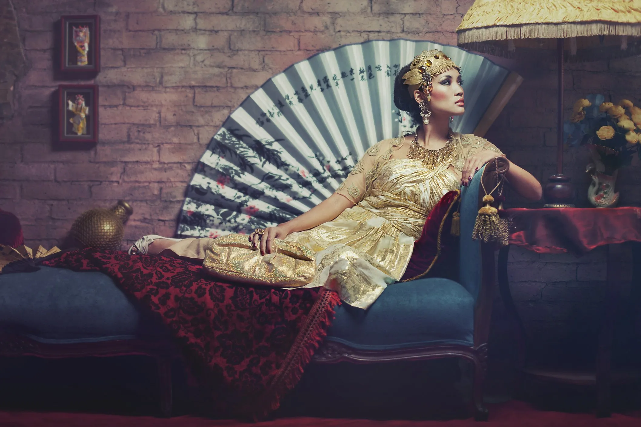 Mujer vestida con ropa dorada y accesorios elaborados, recostada en un sofá azul, con un fondo de pared de ladrillos y abanico decorativo detrás.
