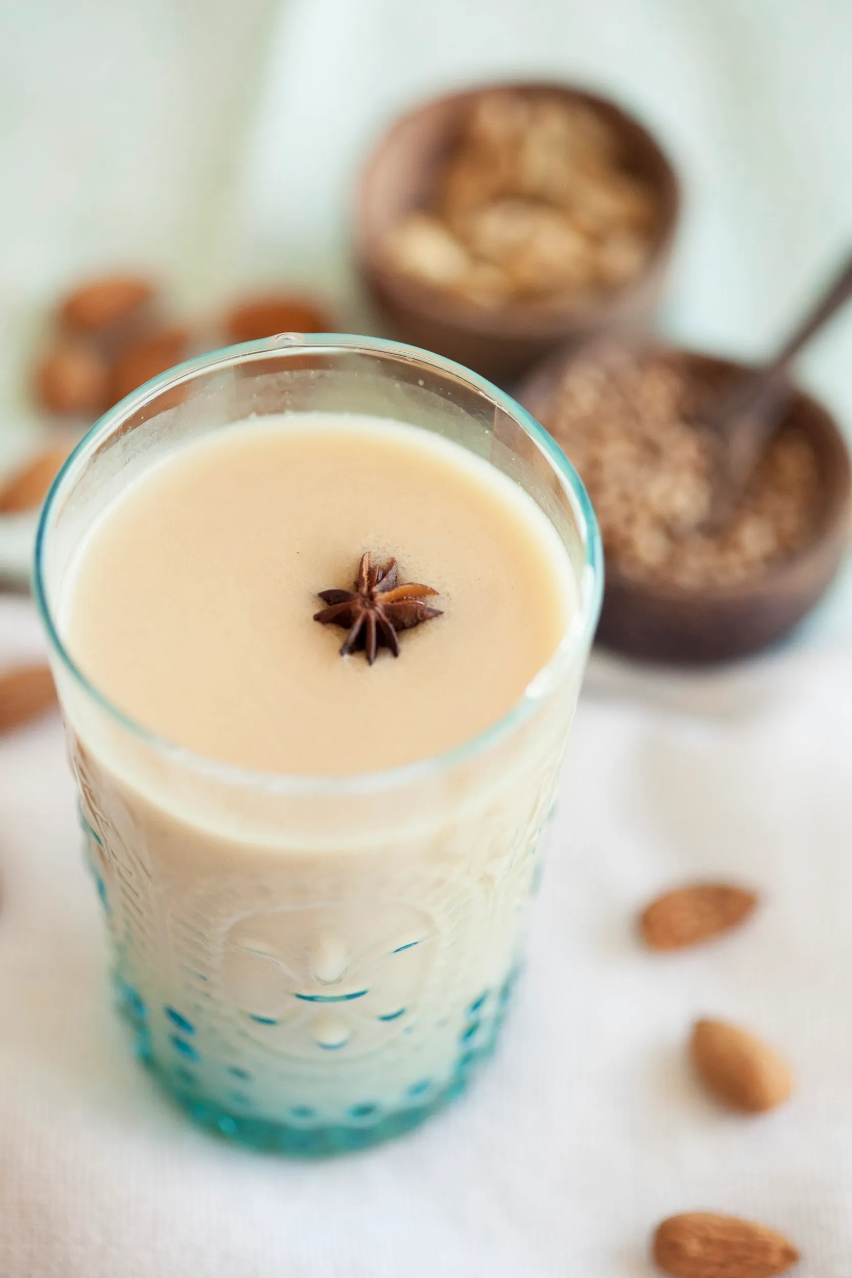 Vaso de leche de almendras con una estrella de anís en la superficie, y en el fondo hay recipientes con almendras, cacao en polvo y azúcar, sobre una mesa blanca.