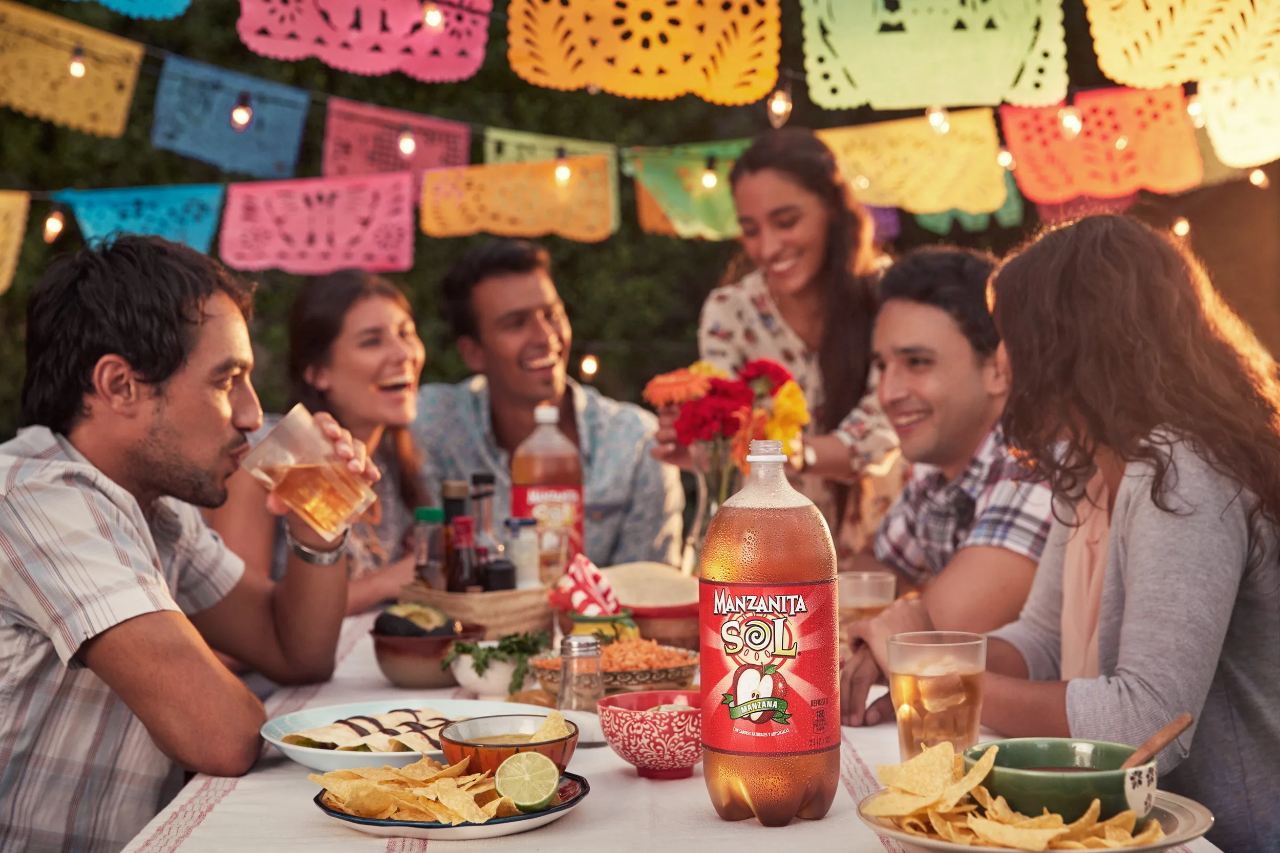 Grupo de personas disfrutando una comida al aire libre, con decoraciones coloridas y una botella de Manzanita Sol en primer plano.