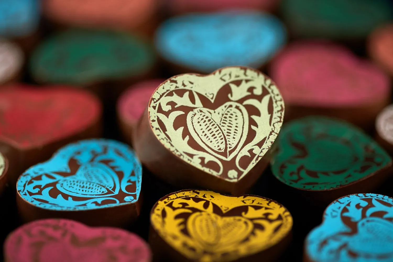 Variados corazones de chocolate con decoraciones coloridas, en primer plano un corazón con diseño ornamental en tonos marrón y crema.
