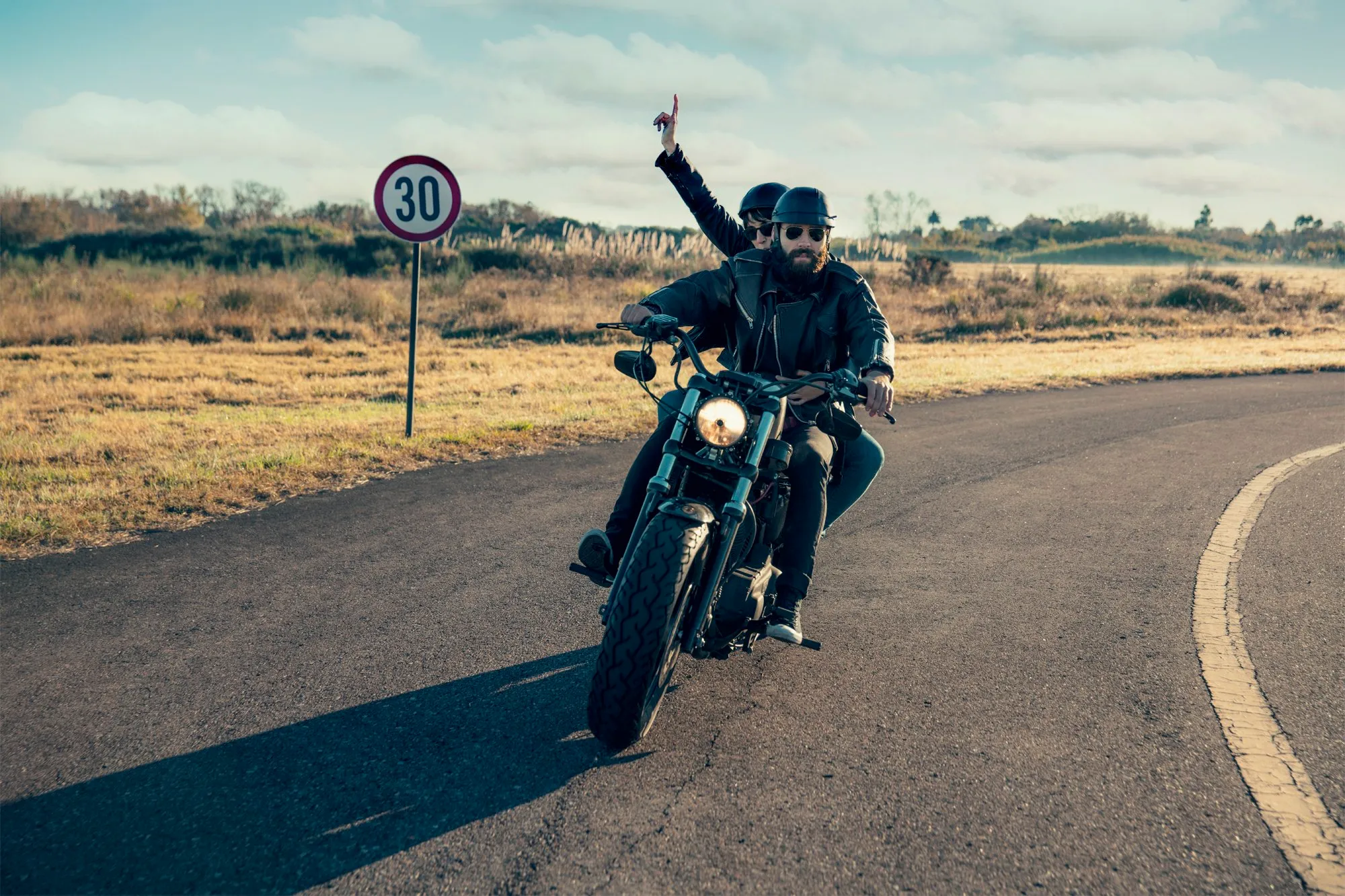 Dos motociclistas con cascos, uno en la parte delantera y otro en la parte trasera, viajando en una motocicleta por una carretera curva en un campo con señal de límite de velocidad de 30 km/h.