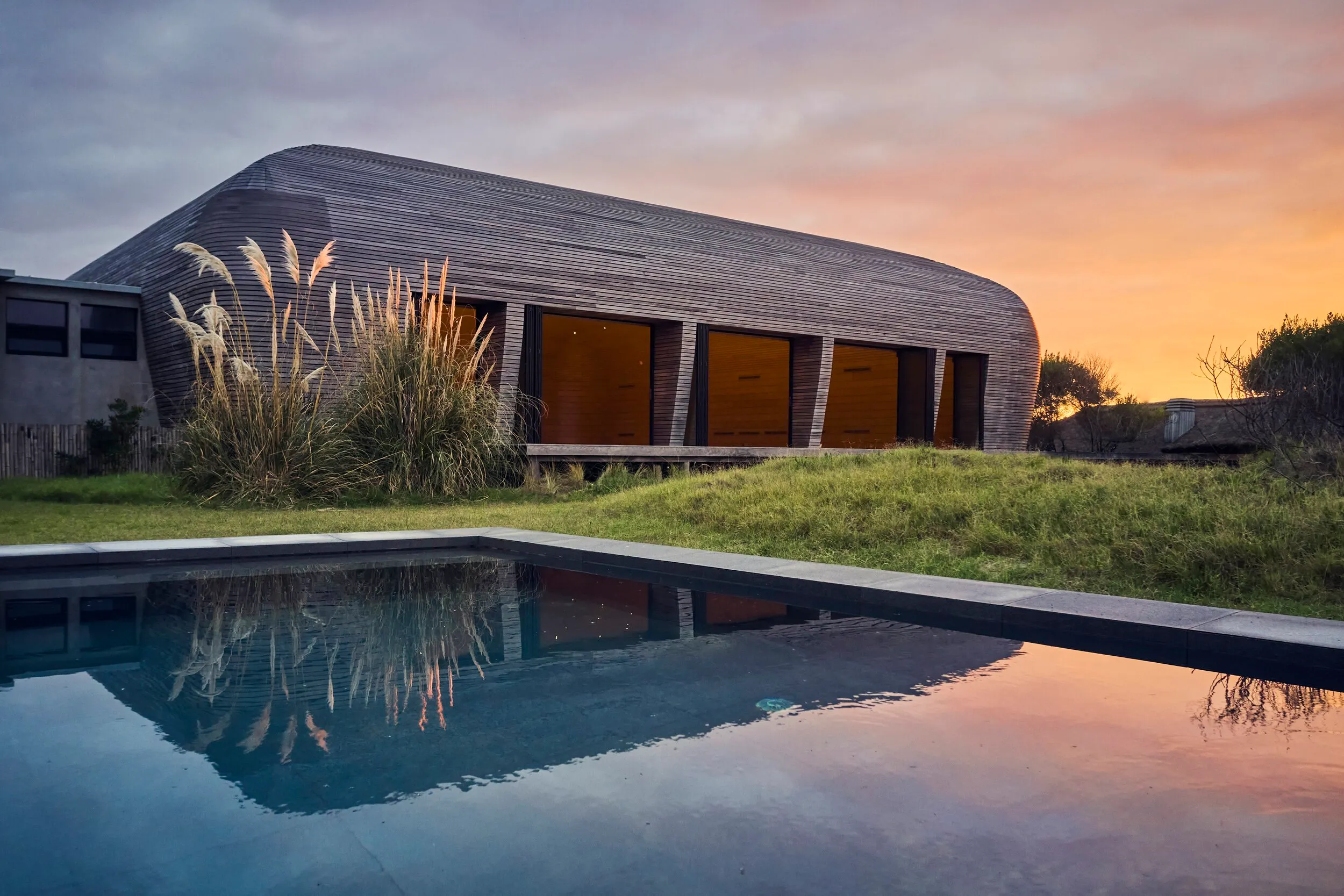 Casa moderna de diseño curvo con grandes ventanas, cerca de una piscina y rodeada de césped, en un atardecer con cielo nublado y tonos cálidos.