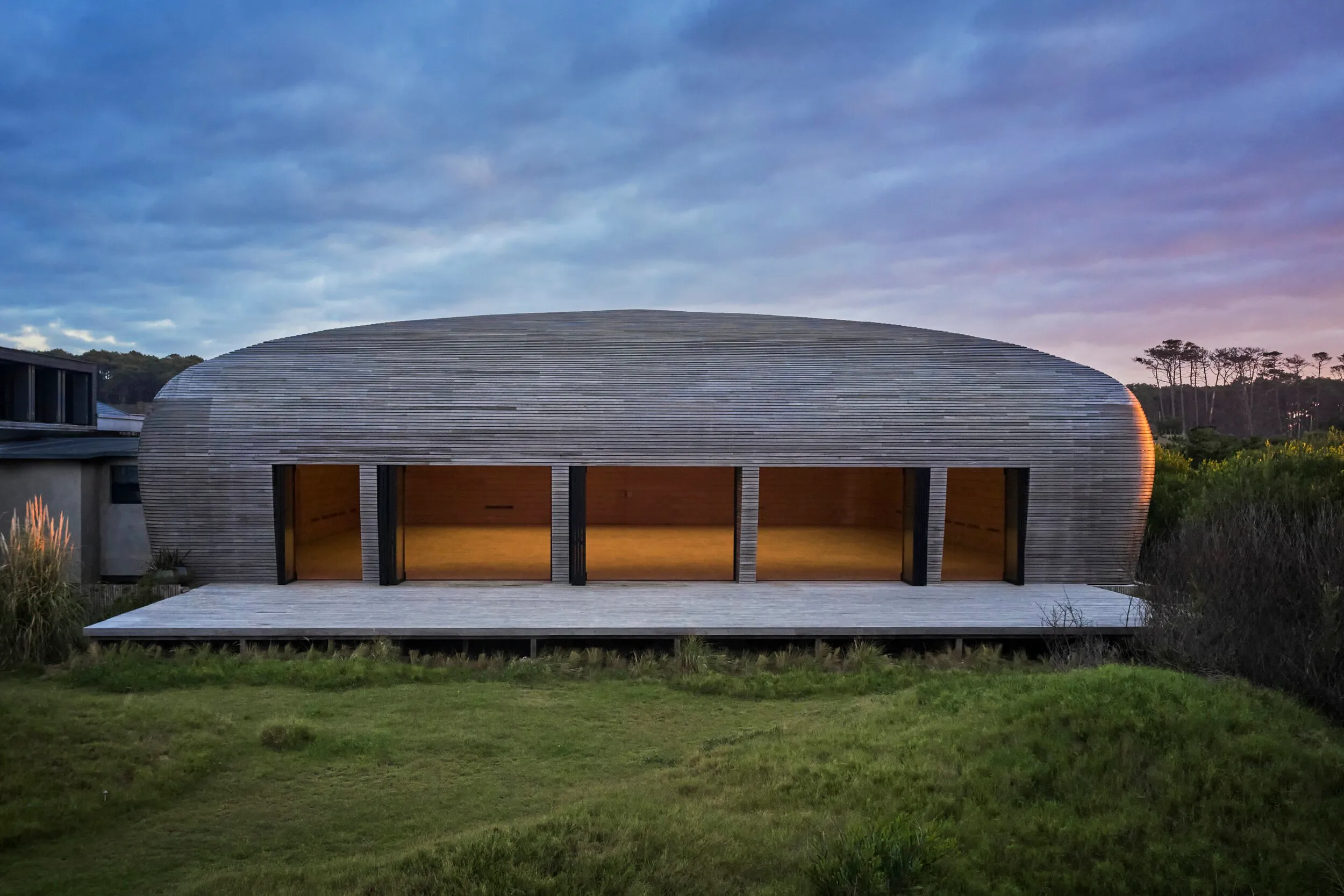 Edificio de diseño moderno con cúpula de madera, grandes ventanas y terraza en un entorno natural al atardecer.