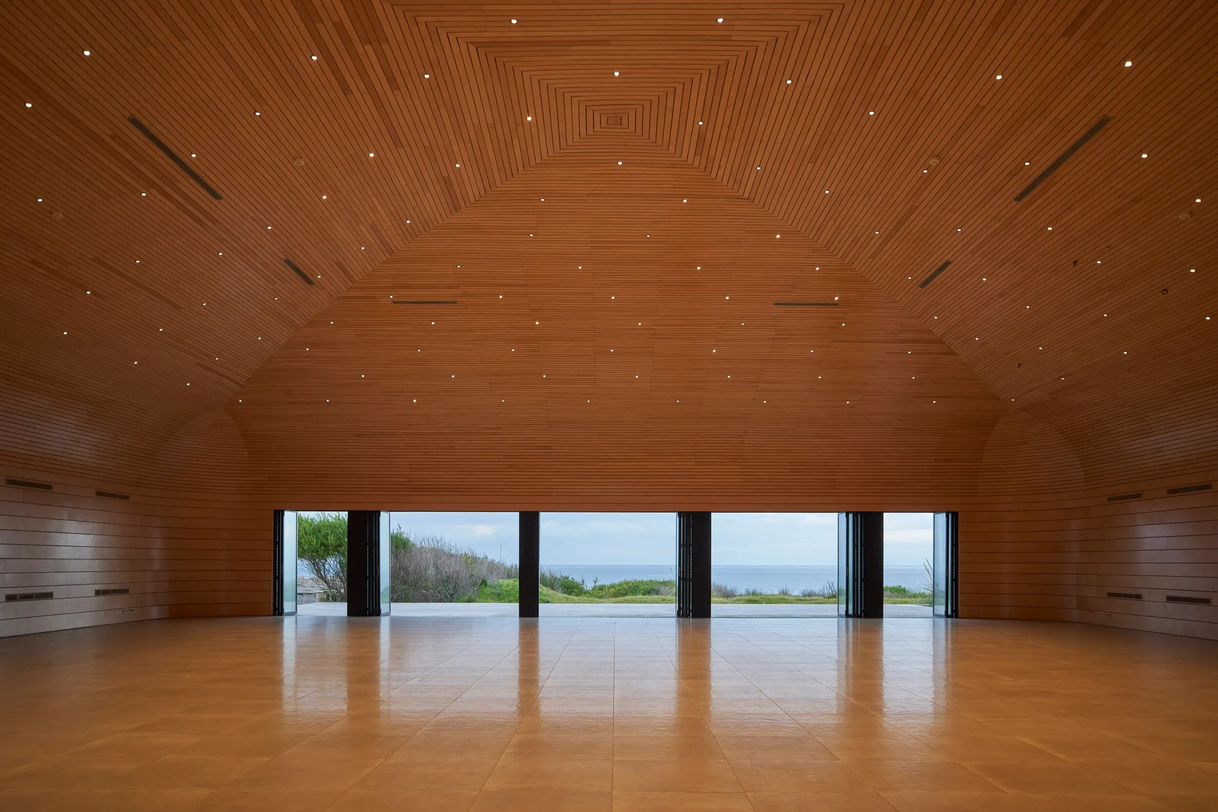 Sala grande con paredes y techo de madera, ventanas abiertas que muestran vista al mar y paisaje con árboles.