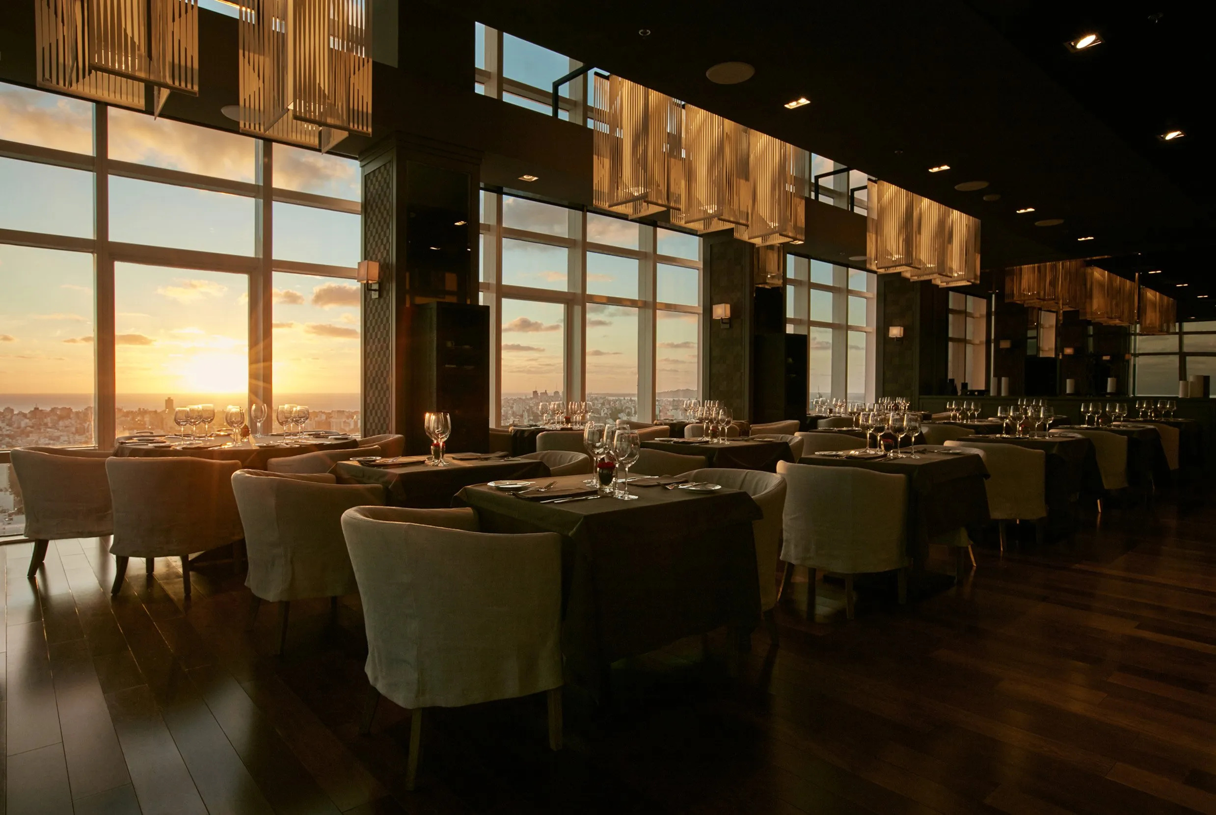 Restaurante elegante con vista al atardecer, mesas con cristalería y cubiertos, en un ambiente oscuro con grandes ventanas y decoraciones modernas.