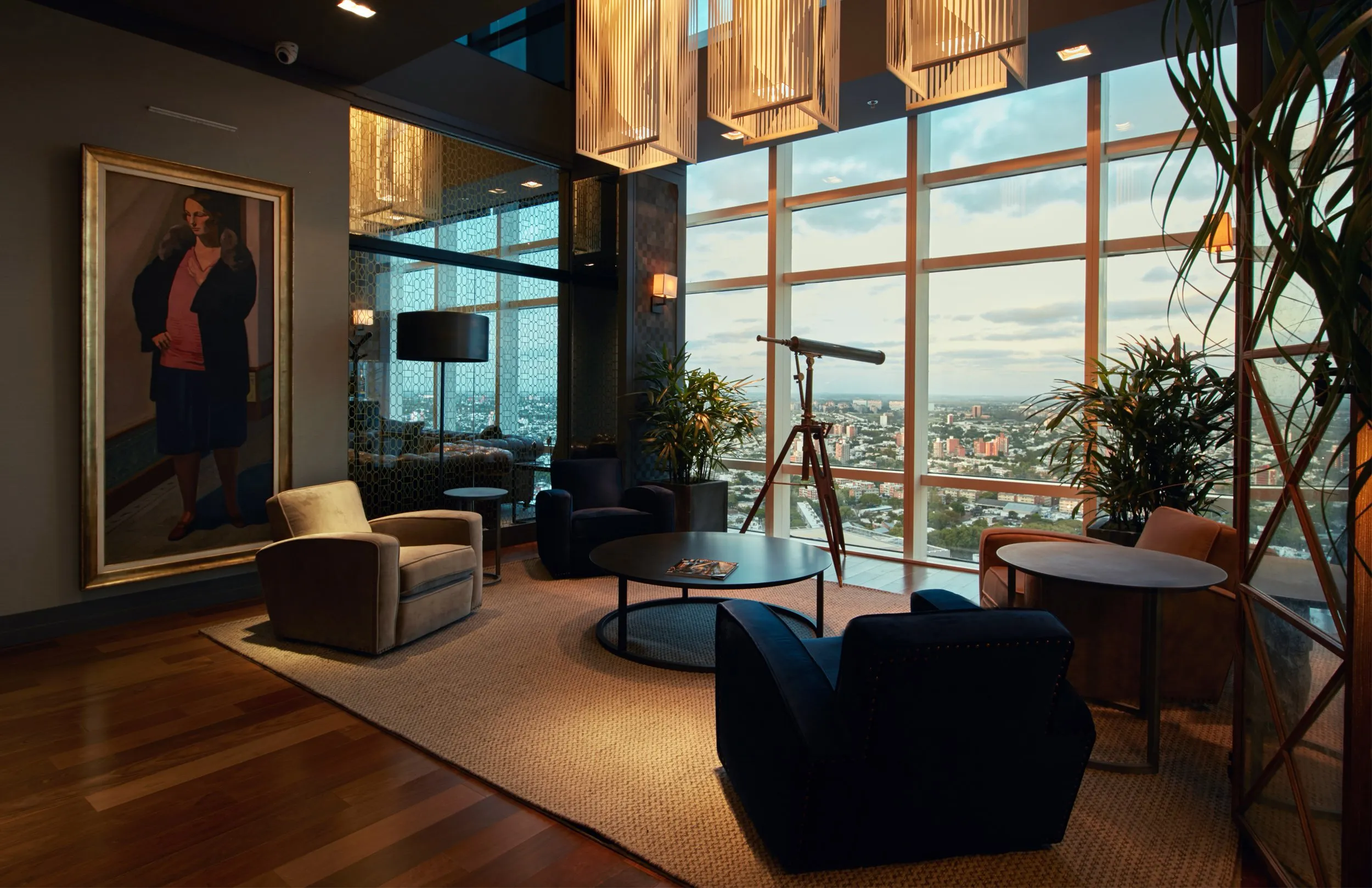 Sala de estar en un edificio alto con vista a la ciudad, sillones y mesa de centro, telescopio junto a grandes ventanas y plantas decorativas.