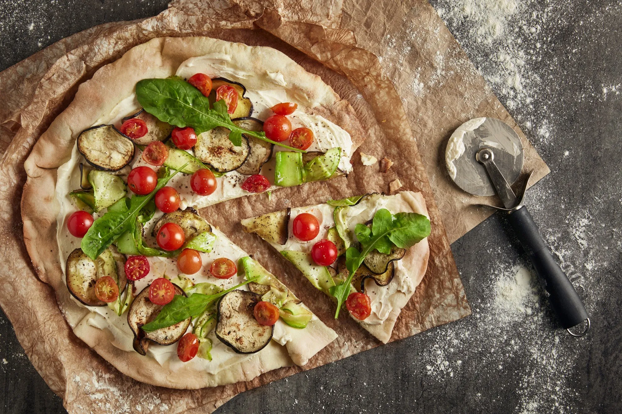Pizza sin hornear con ingredientes como tomate cherry, berenjena, calabacín y hojas de albahaca sobre papel de hornear.