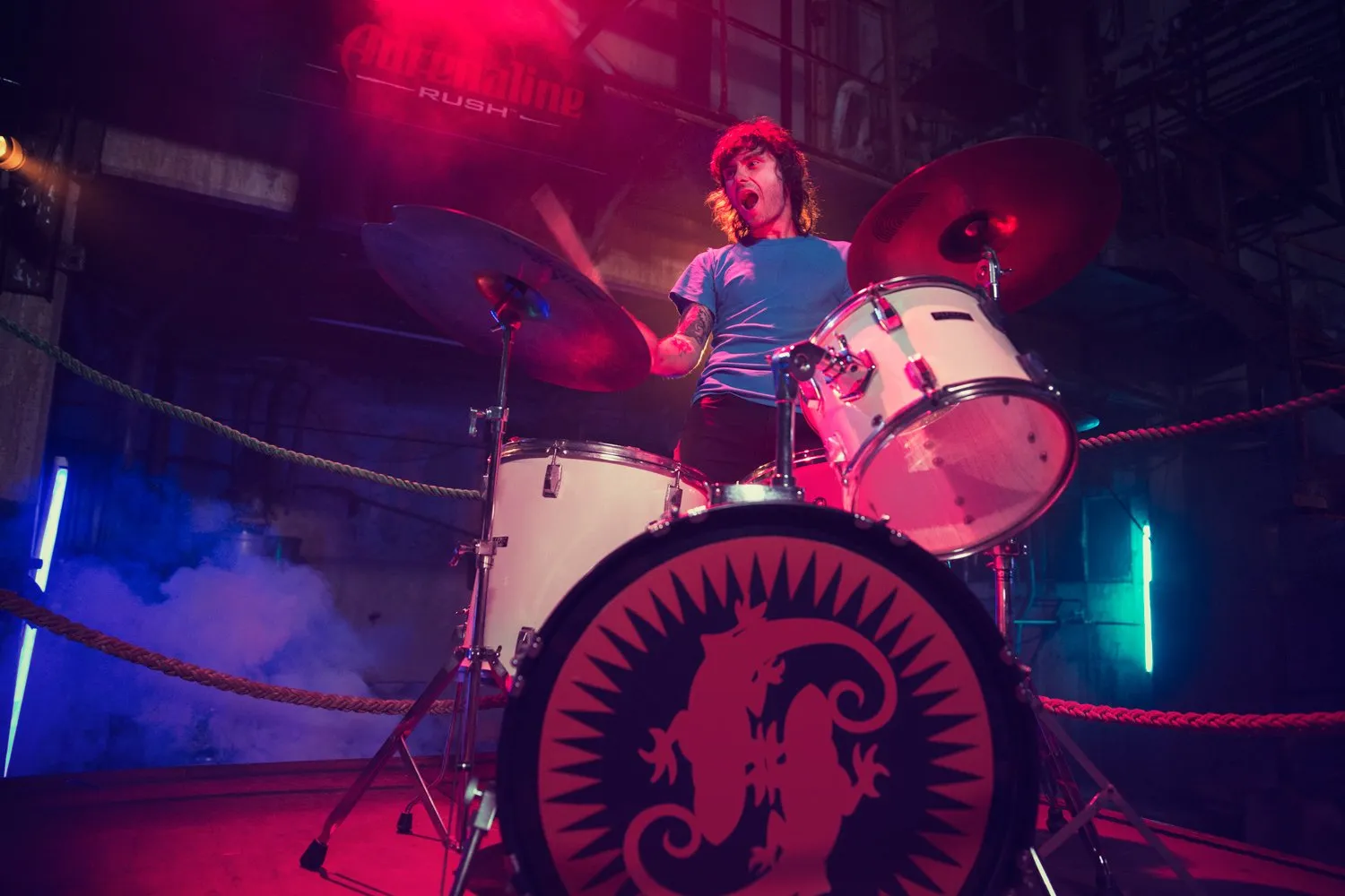 Un hombre tocando la batería en un escenario con luces de colores, aparentemente durante una presentación en vivo.