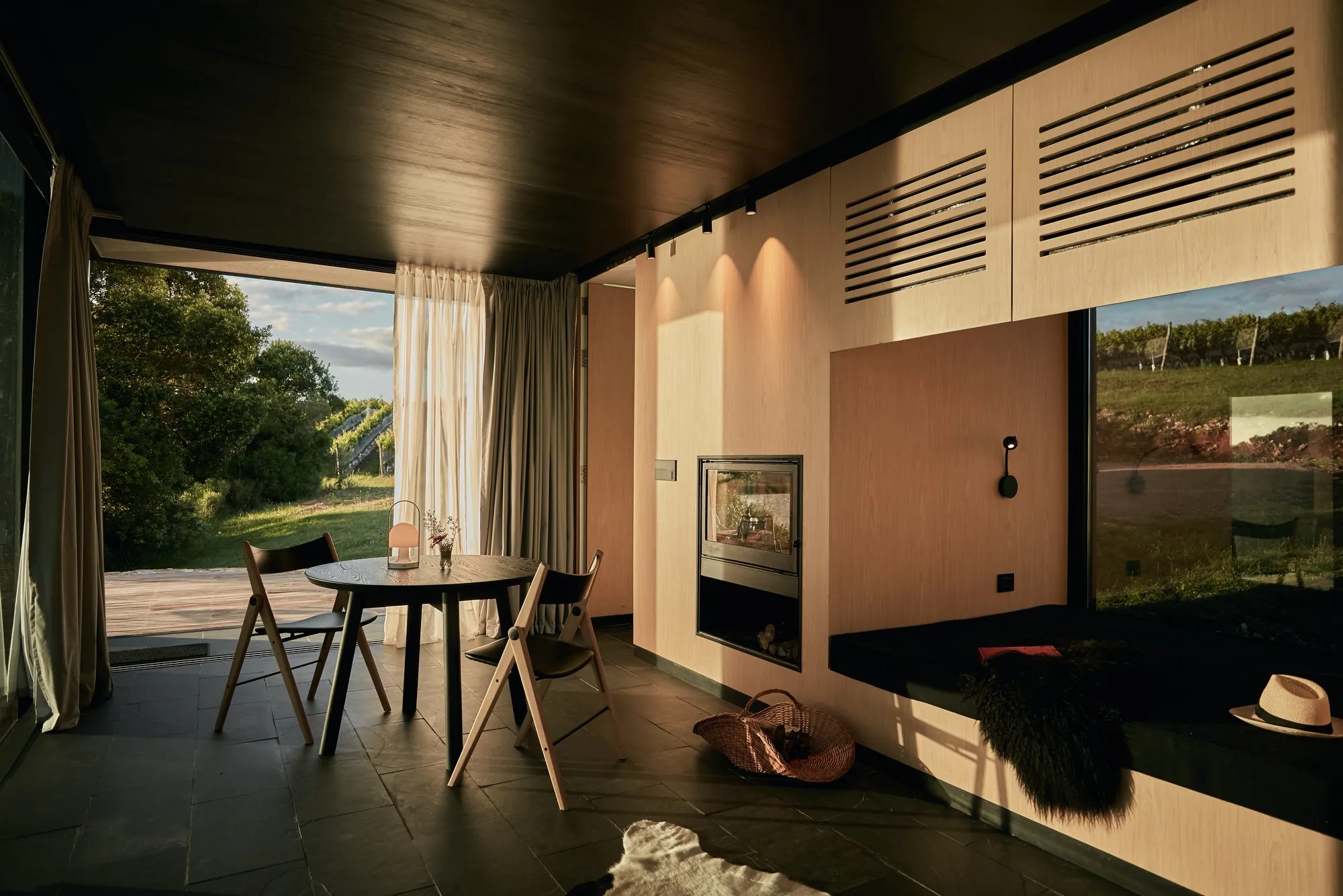 Interior de una estancia moderna con vista a un paisaje natural, mesa redonda con sillas, paredes de madera, chimenea, cortinas blancas y accesorios decorativos