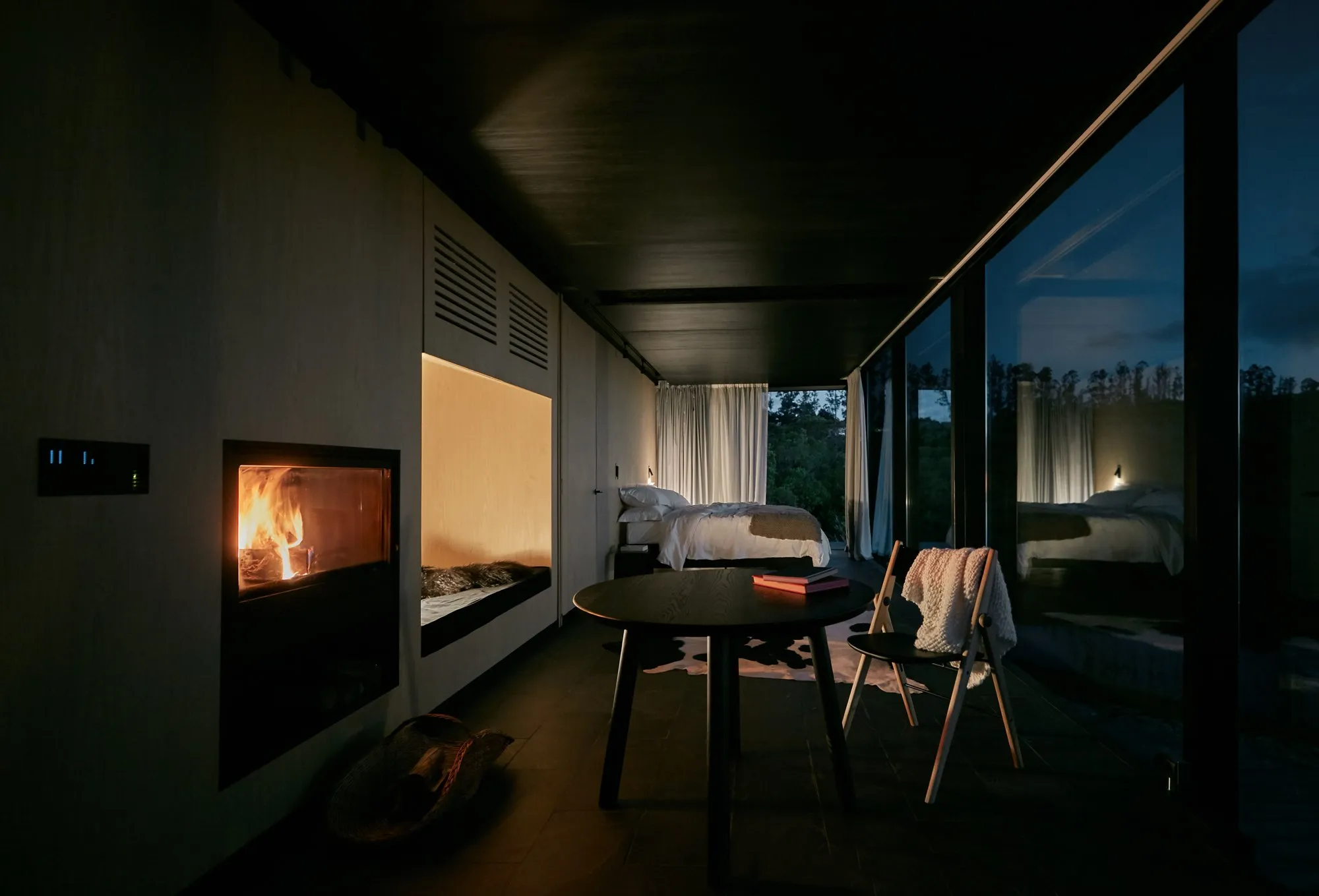 Dormitorio moderno con chimenea encendida, cama grande, vista a la naturaleza a través de grandes ventanas y cortinas blancas, decoración minimalista.