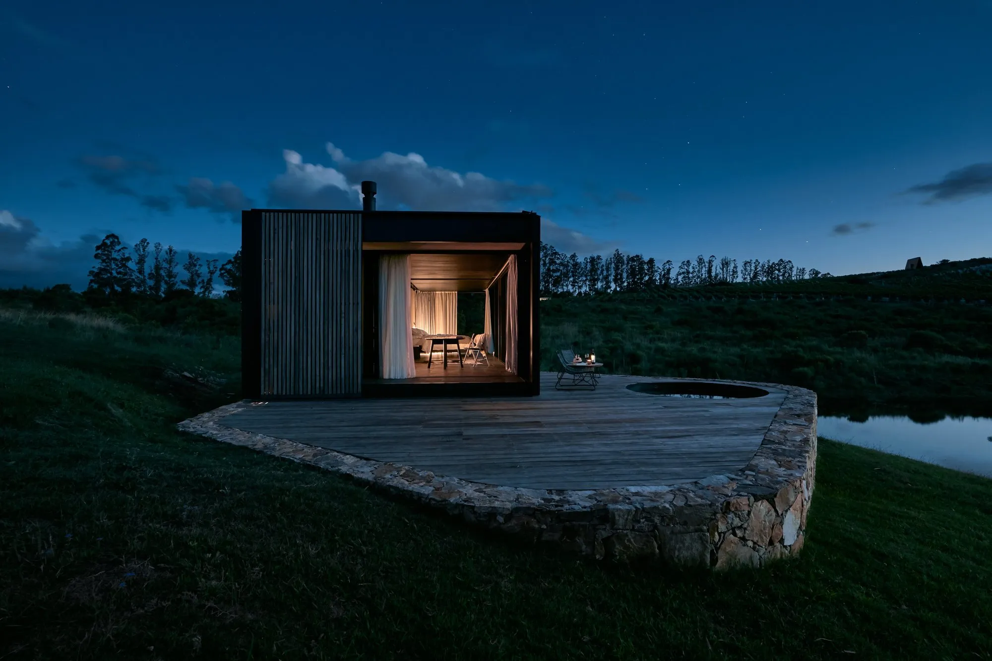 Casa moderna de estructura rectangular con paredes de madera, ventanas con cortinas blancas, en un entorno natural con césped y un pequeño cuerpo de agua al lado, durante la noche con cielo estrellado.