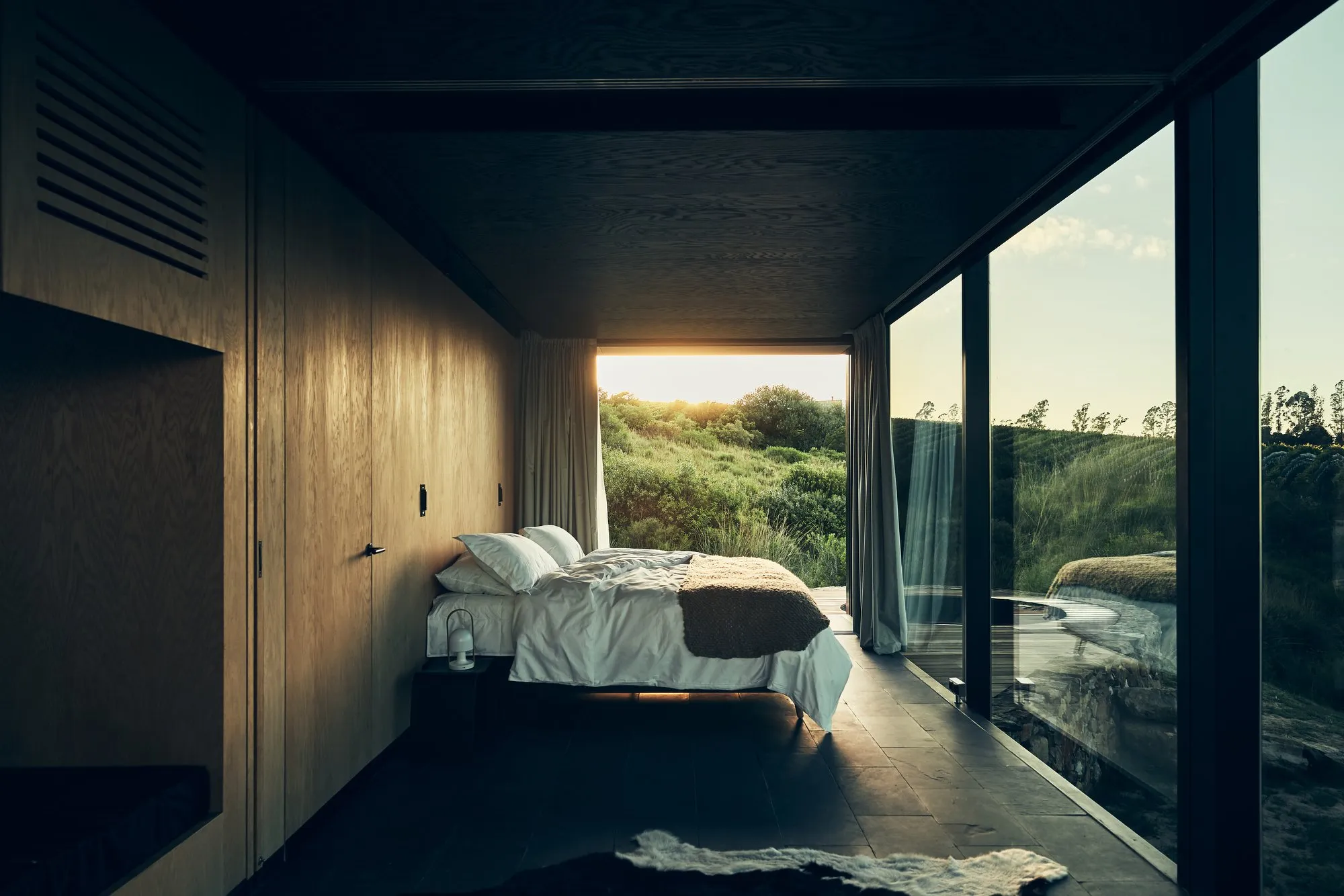 Habitación moderna con vista a la naturaleza, con cama, paredes de madera, y grandes ventanales con cortinas blancas que dejan ver un paisaje verde y soleado.