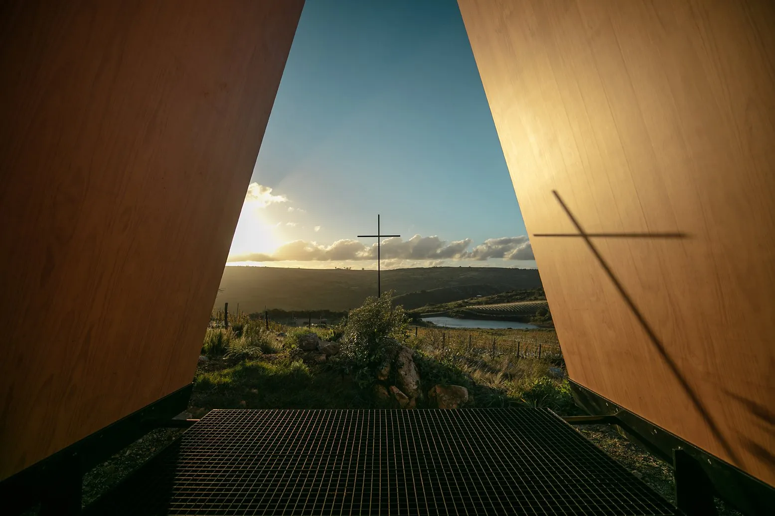 Vista de un paisaje campestre con colinas, un río y un crucifijo en el fondo, vista desde una estructura de madera con marco de metal en primer plano, durante el atardecer.