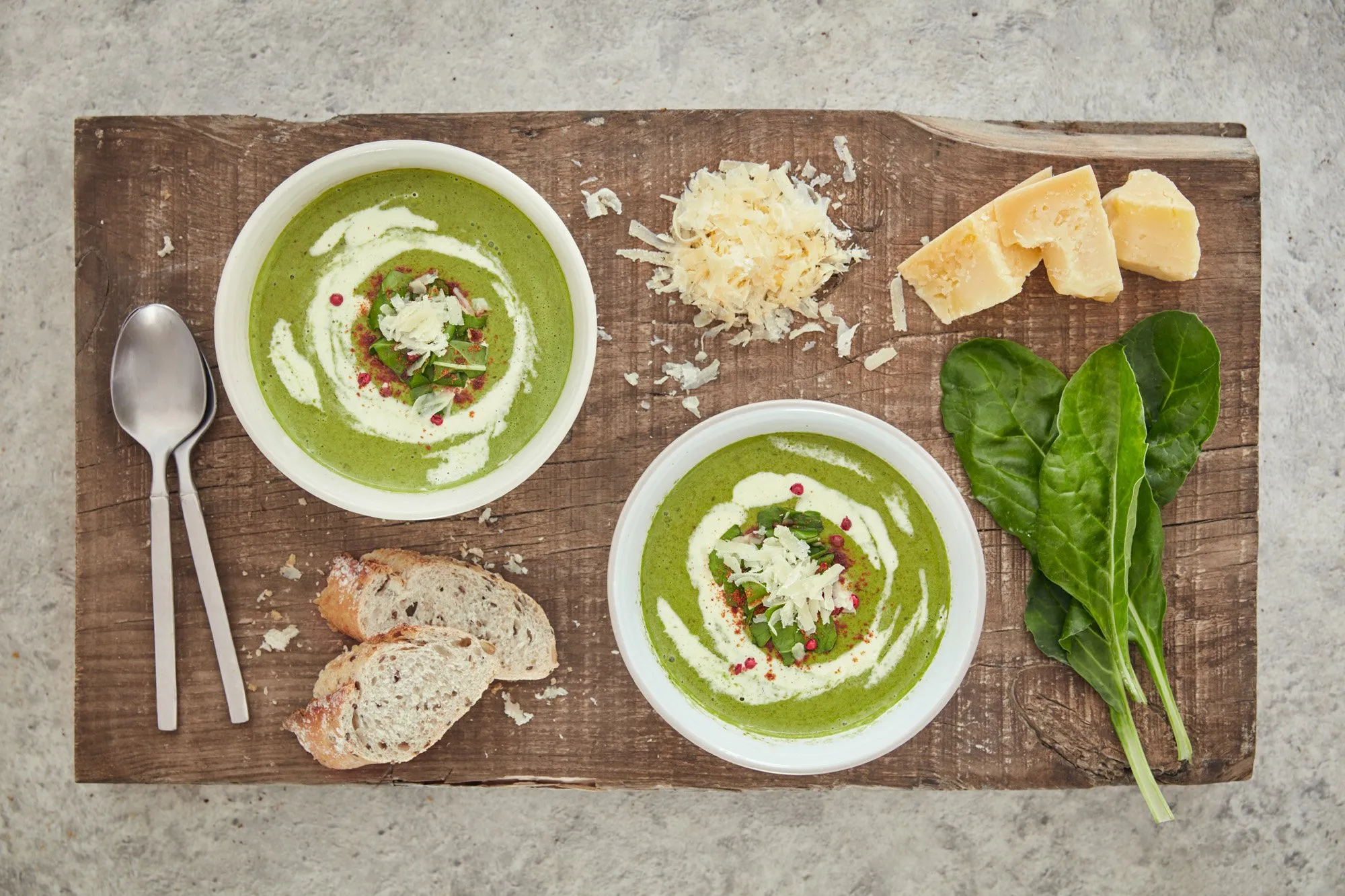 Sopa de espinaca con crema y queso rallado en platos blancos, pan casero, queso, espinaca y hierbas en una tabla de madera.