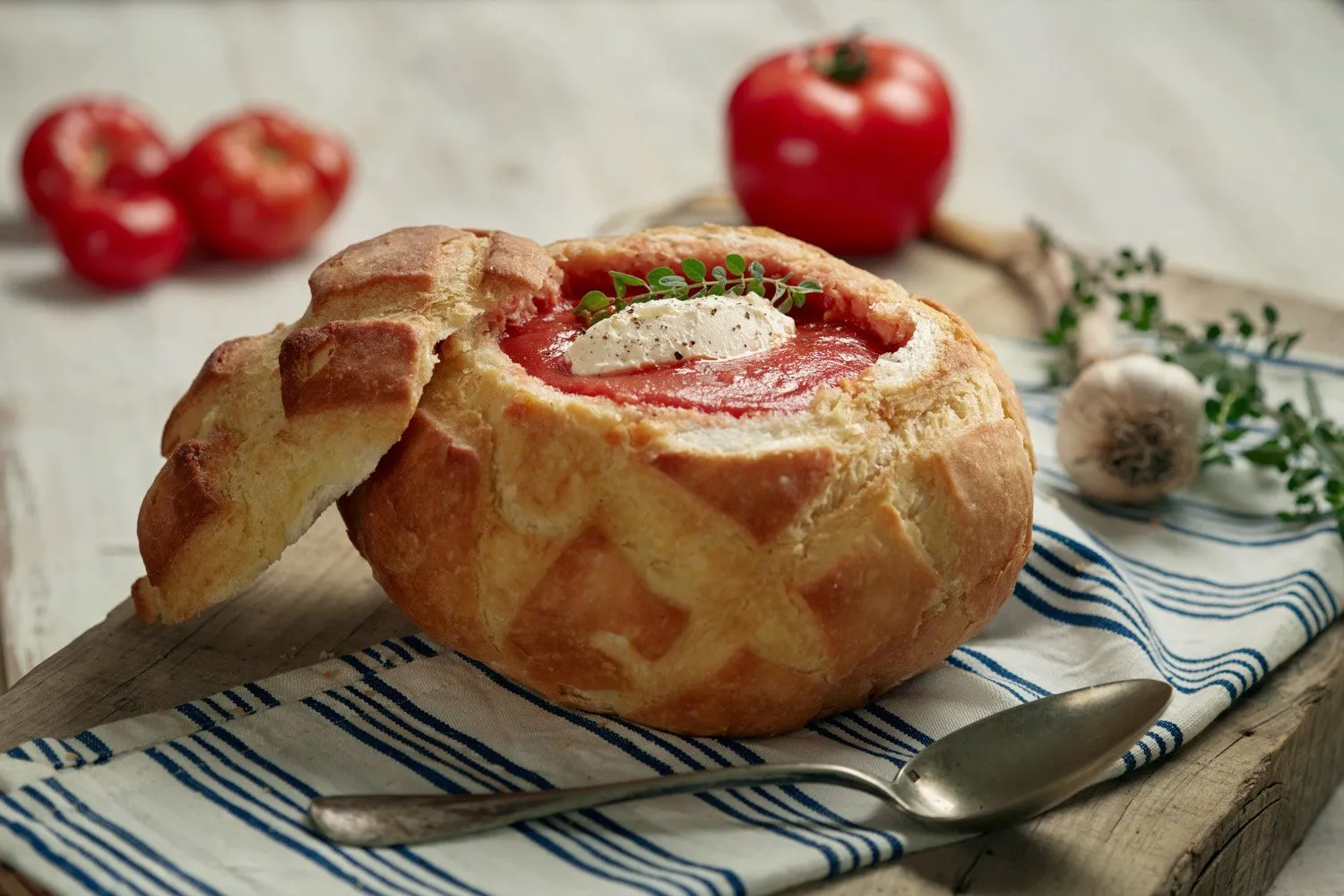 Una sopa en un pan horneado, con queso, tomate y hierbas, sobre una tabla de madera con una cuchara y tomates en el fondo.