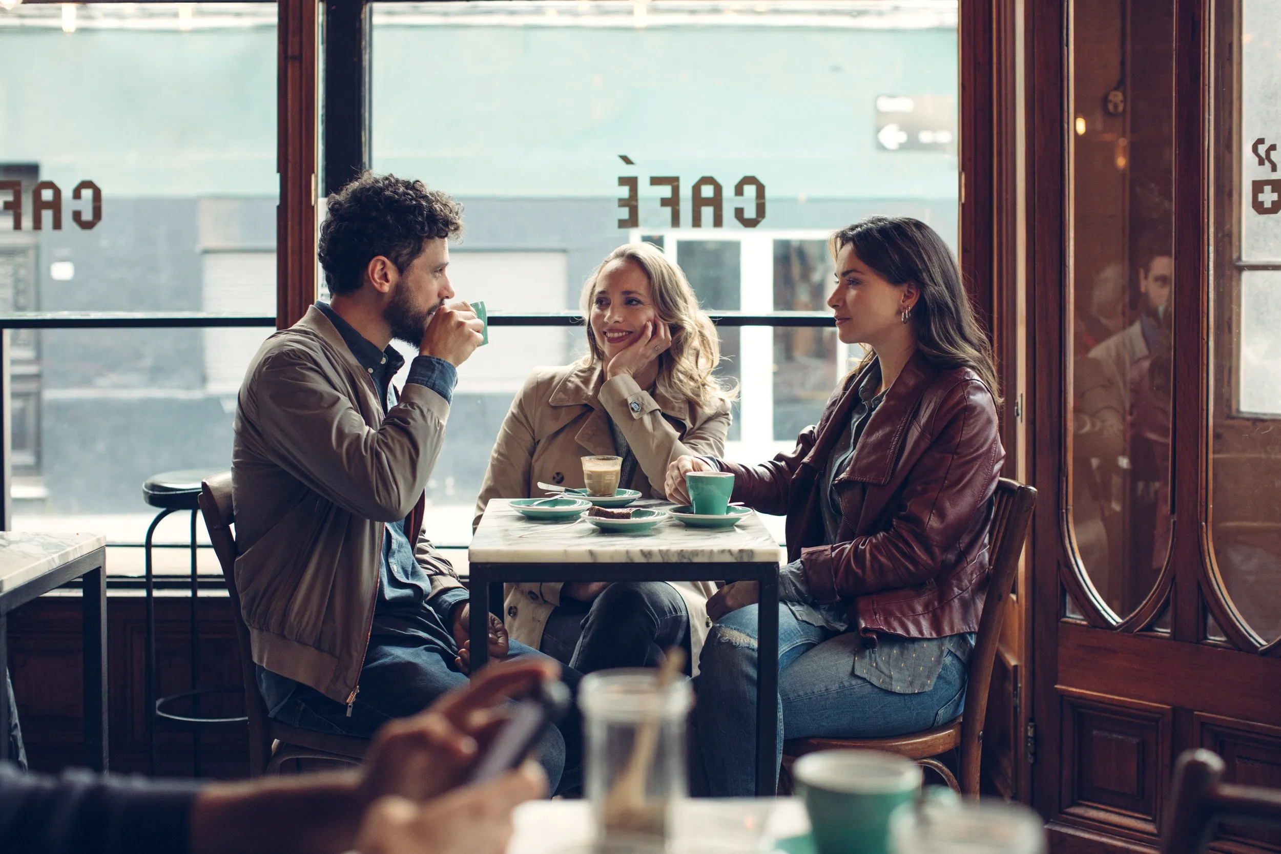 Tres personas sentadas en una mesa de cafetería, conversando y tomando café.