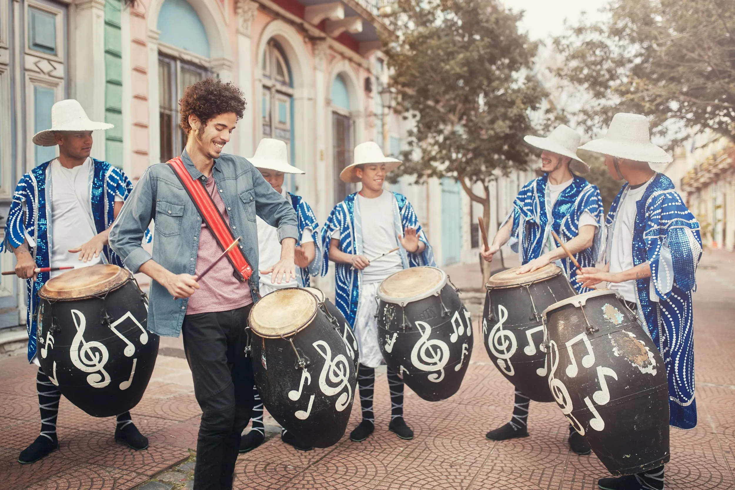 Grupo de músicos tocando tambores decorados con notas musicales en una calle con árboles y edificios coloridos.