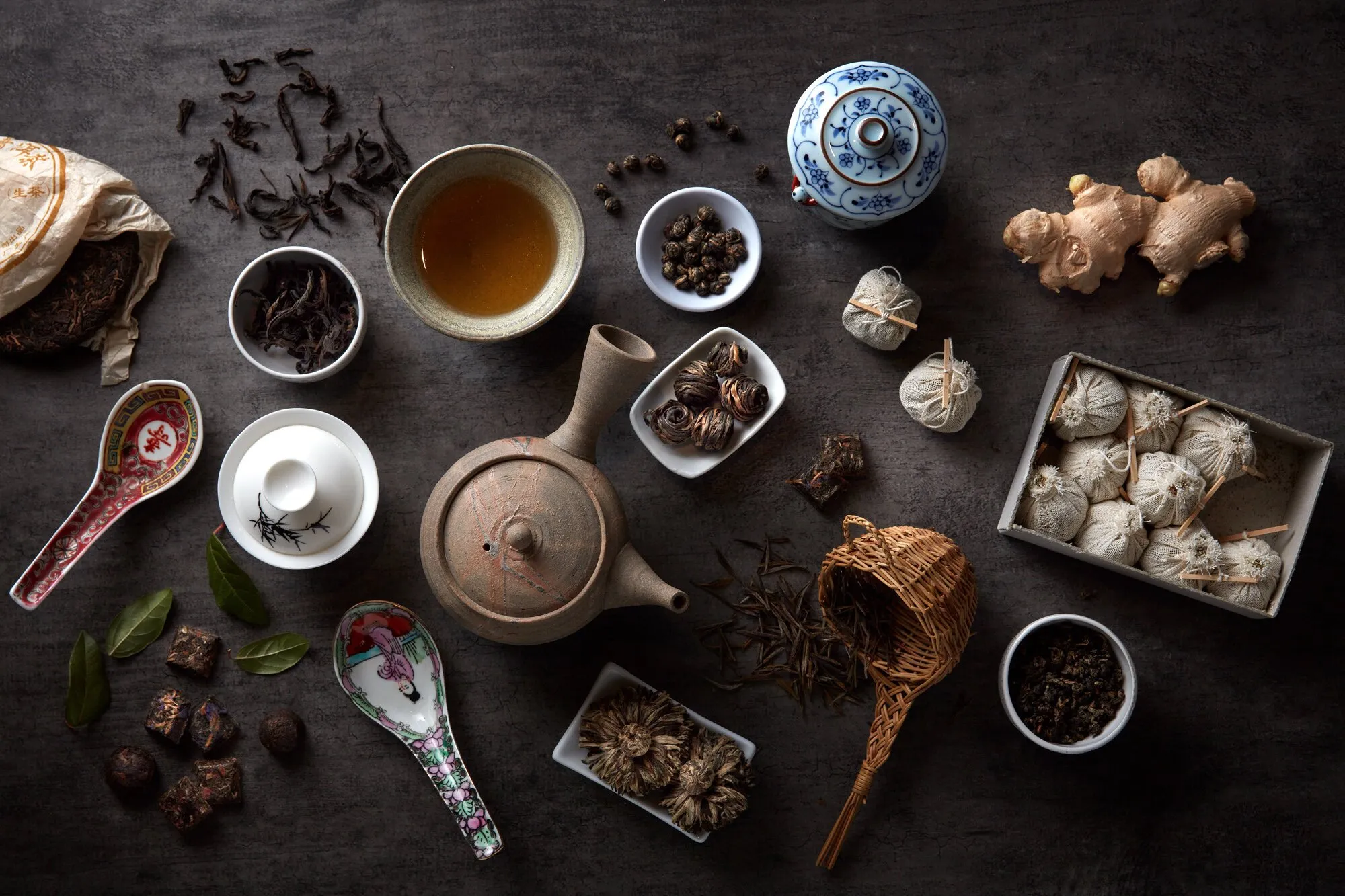 Diversos ingredientes y utensilios para preparar té chino, incluyendo tés sueltos, jengibre, y accesorios tradicionales en una superficie de madera oscura.