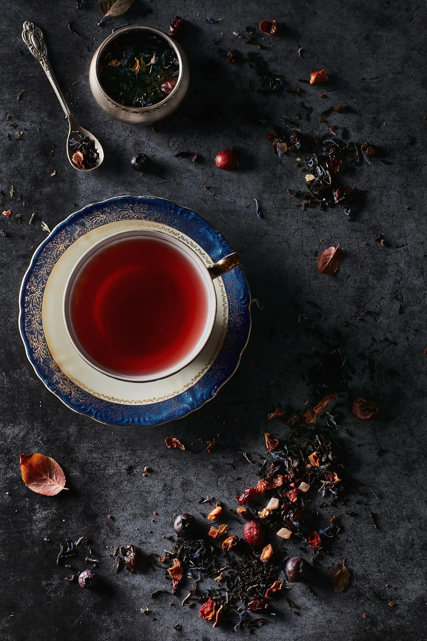 Una taza de té rojo en un platillo decorativo azul y dorado, rodeada de flores secas, hojas y pedazos de fruta que parecen ser arándanos y gajos de pétalos de rosa, sobre una superficie oscura.