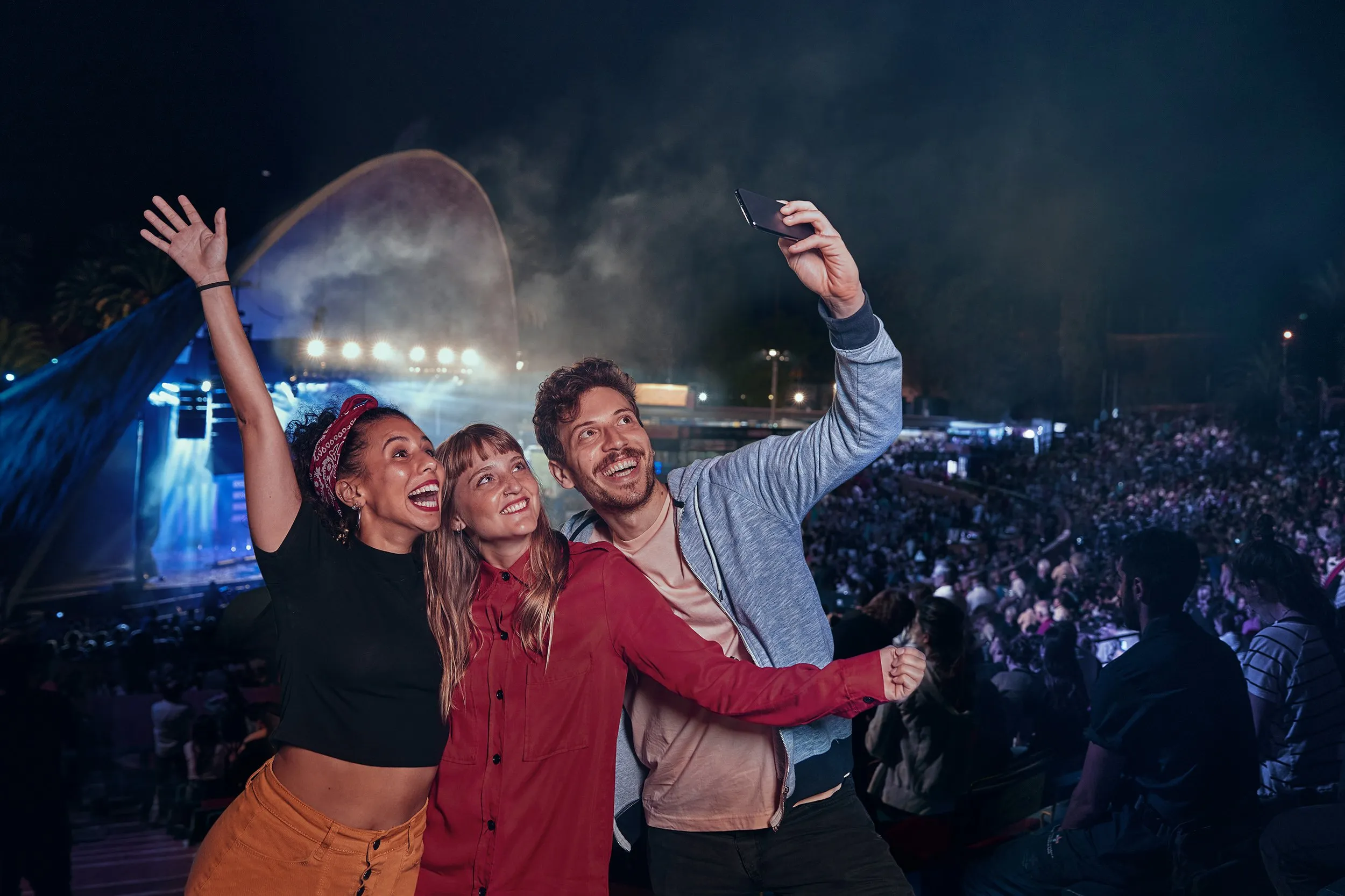 Tres personas felices tomándose una selfie en un concierto al aire libre en la noche, con una audiencia y un escenario iluminado al fondo.
