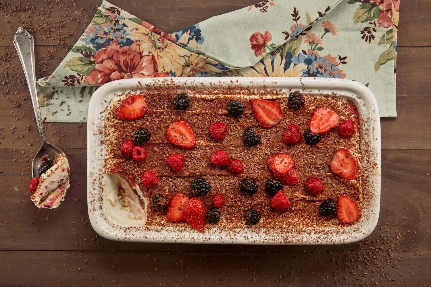 Postre de tiramisú cubierto con frambuesas, fresas y moras, en un plato rectangular blanco sobre una mesa de madera. Un tenedor con un trozo del postre y una servilleta con un diseño floral están a la izquierda.
