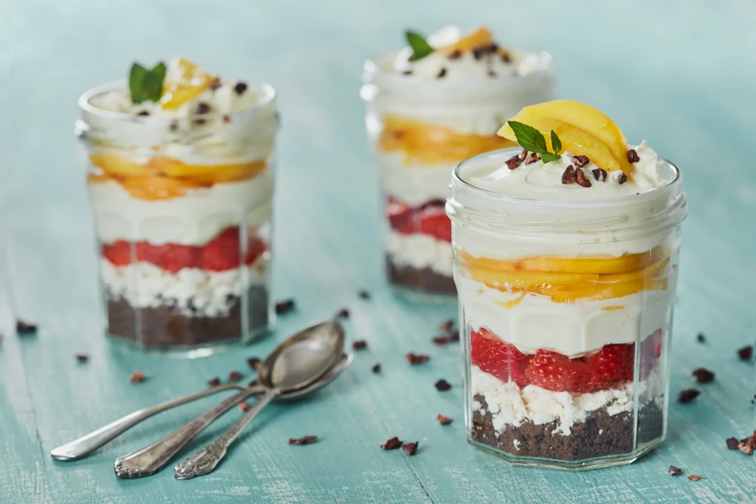 Copas con capas de pastel de chocolate, crema, fresas, mango y decoradas con menta y cacao en virutas sobre una mesa de madera azul.
