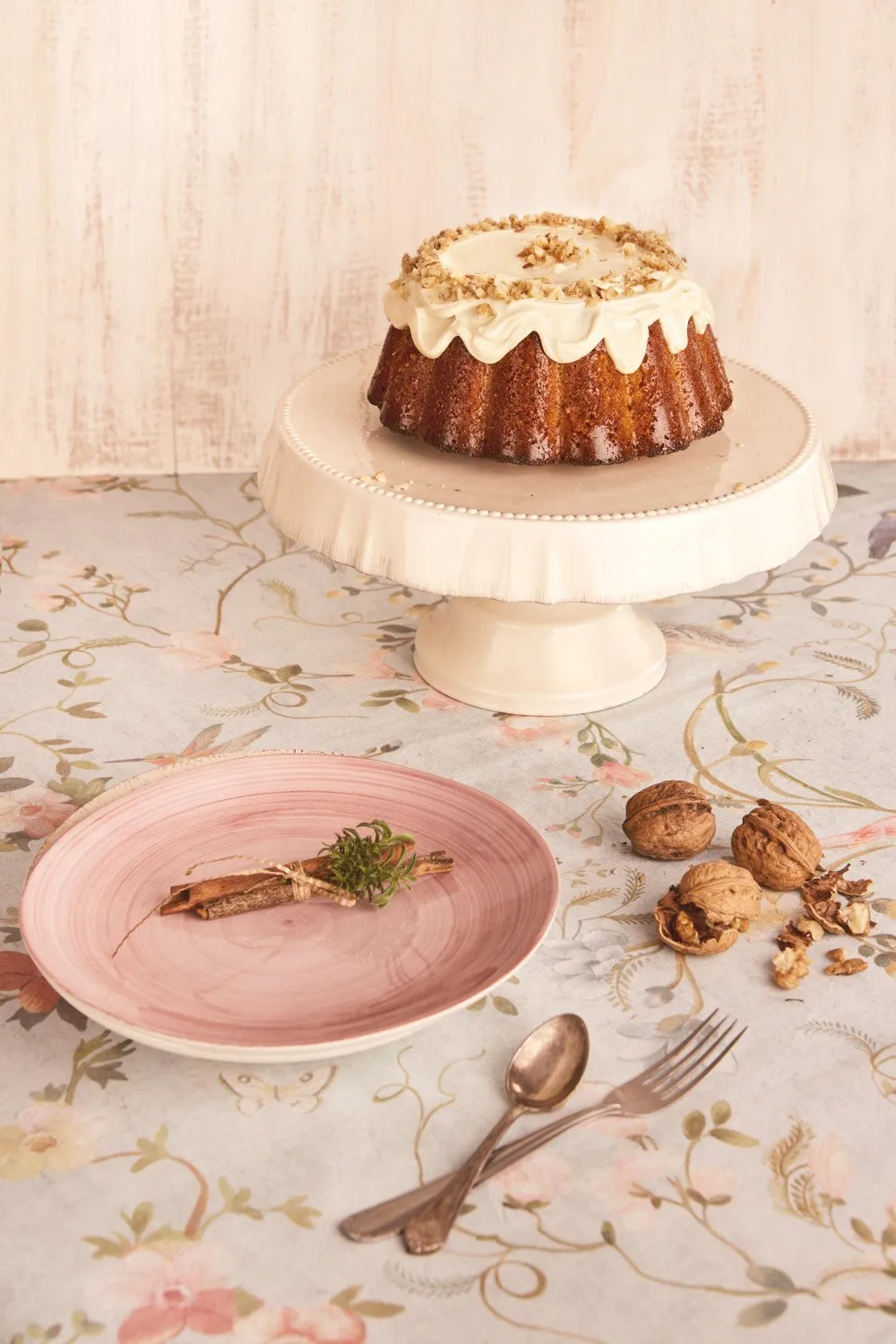 Pastel de amado en soporte con decoraciones de nueces y crema, plato rosa con ramito de plantas y nueces en la mesa, cubiertos de plata junto a un fondo de flores en la mesa