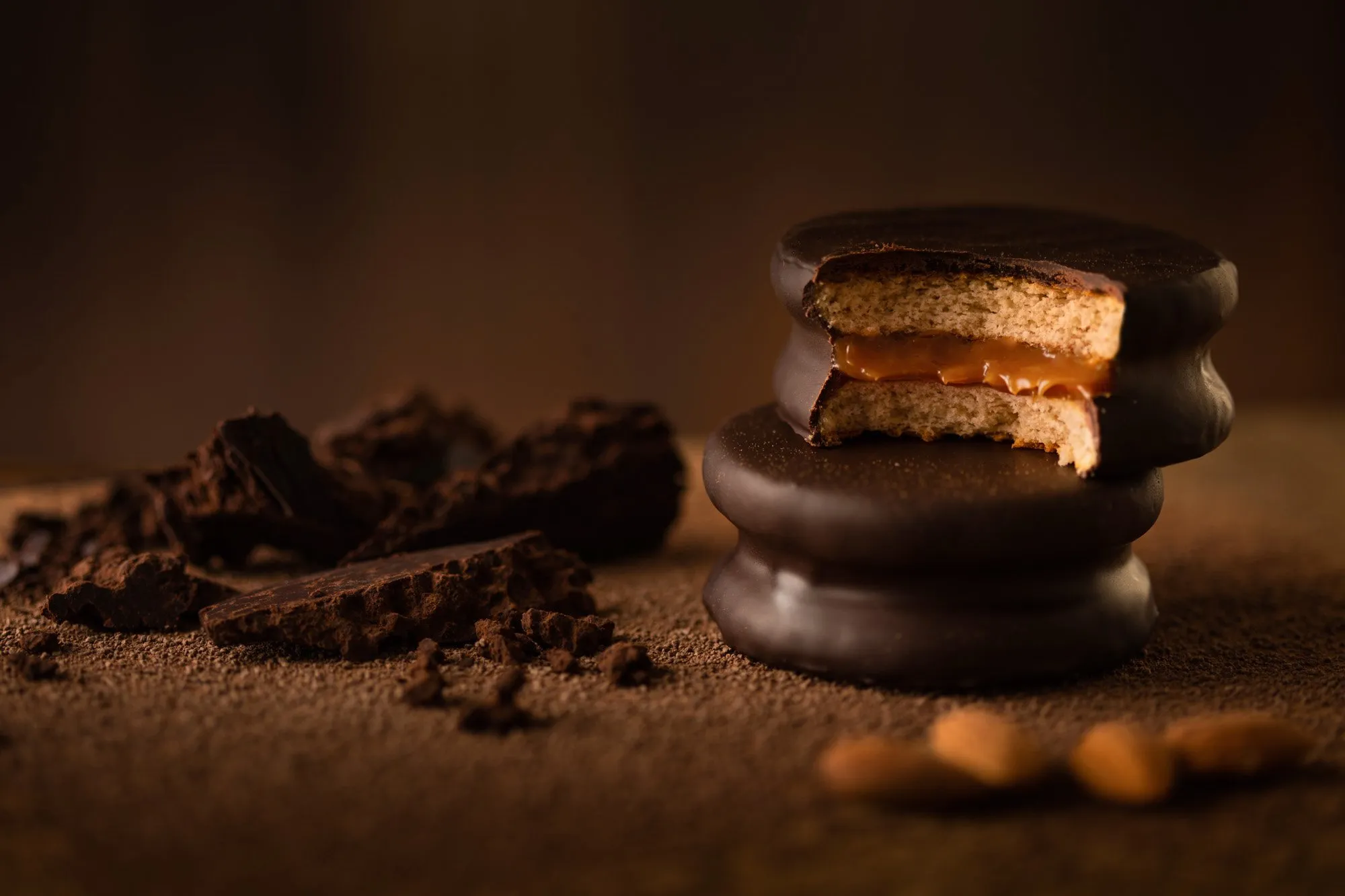 Galleta con relleno de caramelo, cubierta de chocolate, sobre una superficie de cacao en polvo junto a trozos de chocolate