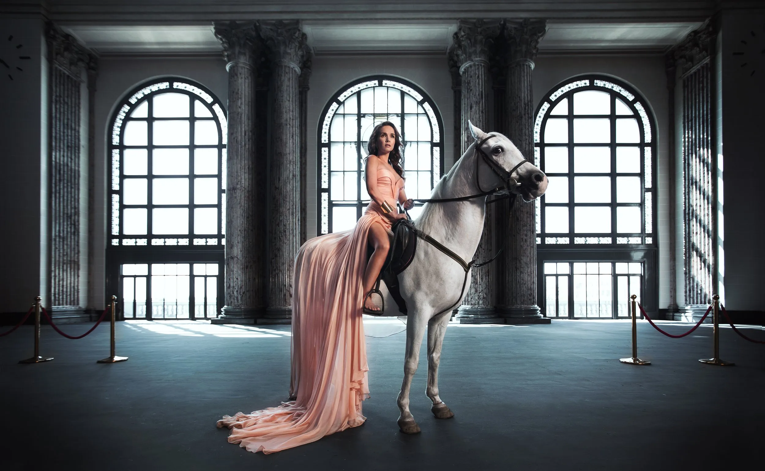Mujer con vestido largo en tono rosa claro montada en un caballo blanco en un interior con columnas de piedra y ventanas altas