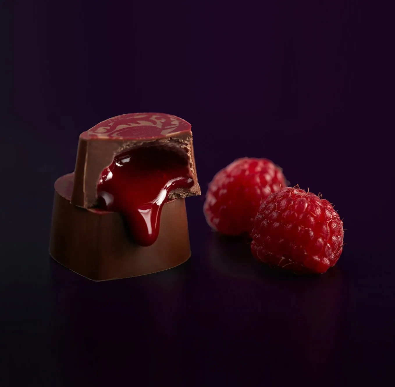Chocolate relleno de salsa de frambuesa junto a dos frambuesas frescas.