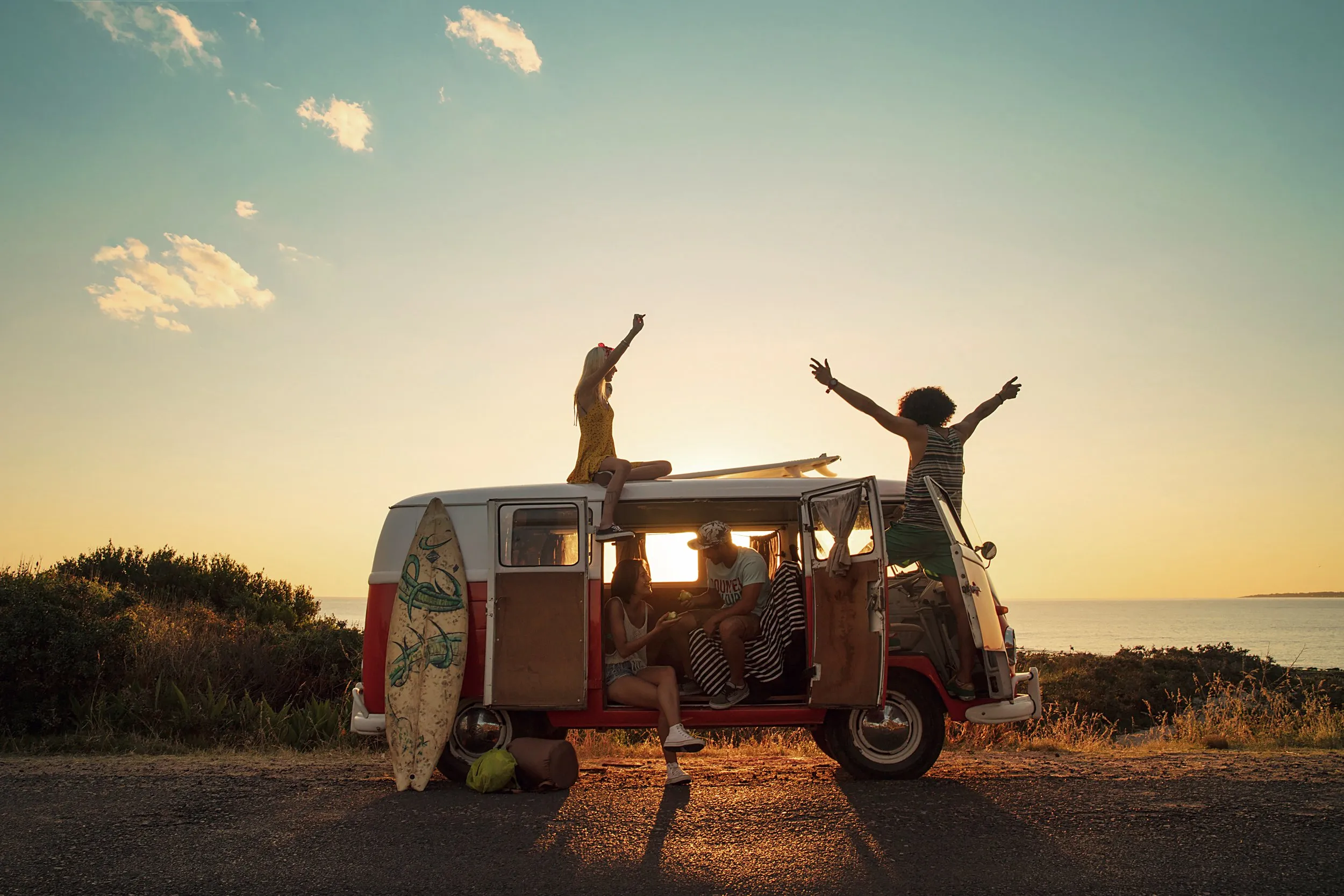 Grupo de amigos disfrutando en una furgoneta con vista al mar al atardecer, con una chica en la azotea y otras dentro, en un ambiente relajado y alegre.