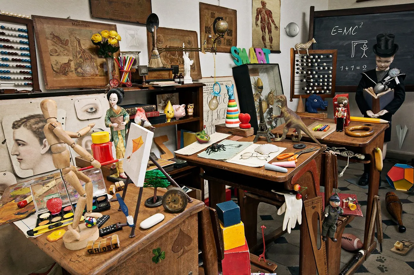 Una sala de clases llena de objetos diversos, como marionetas, libros, juguetes, instrumentos científicos y artículos de arte, con varias mesas y paredes decoradas con cuadros, dibujos y herramientas educativas.