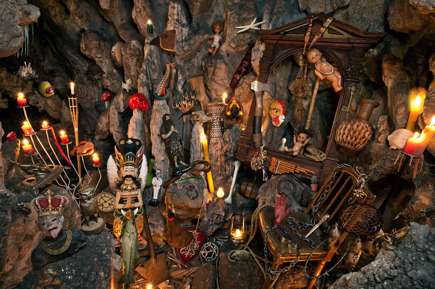Un altar con objetos rituales y decorativos, incluyendo velas encendidas, máscaras, figuras, armas, cadenas, un pescado colgado y otros elementos diversos en un entorno de piedra.